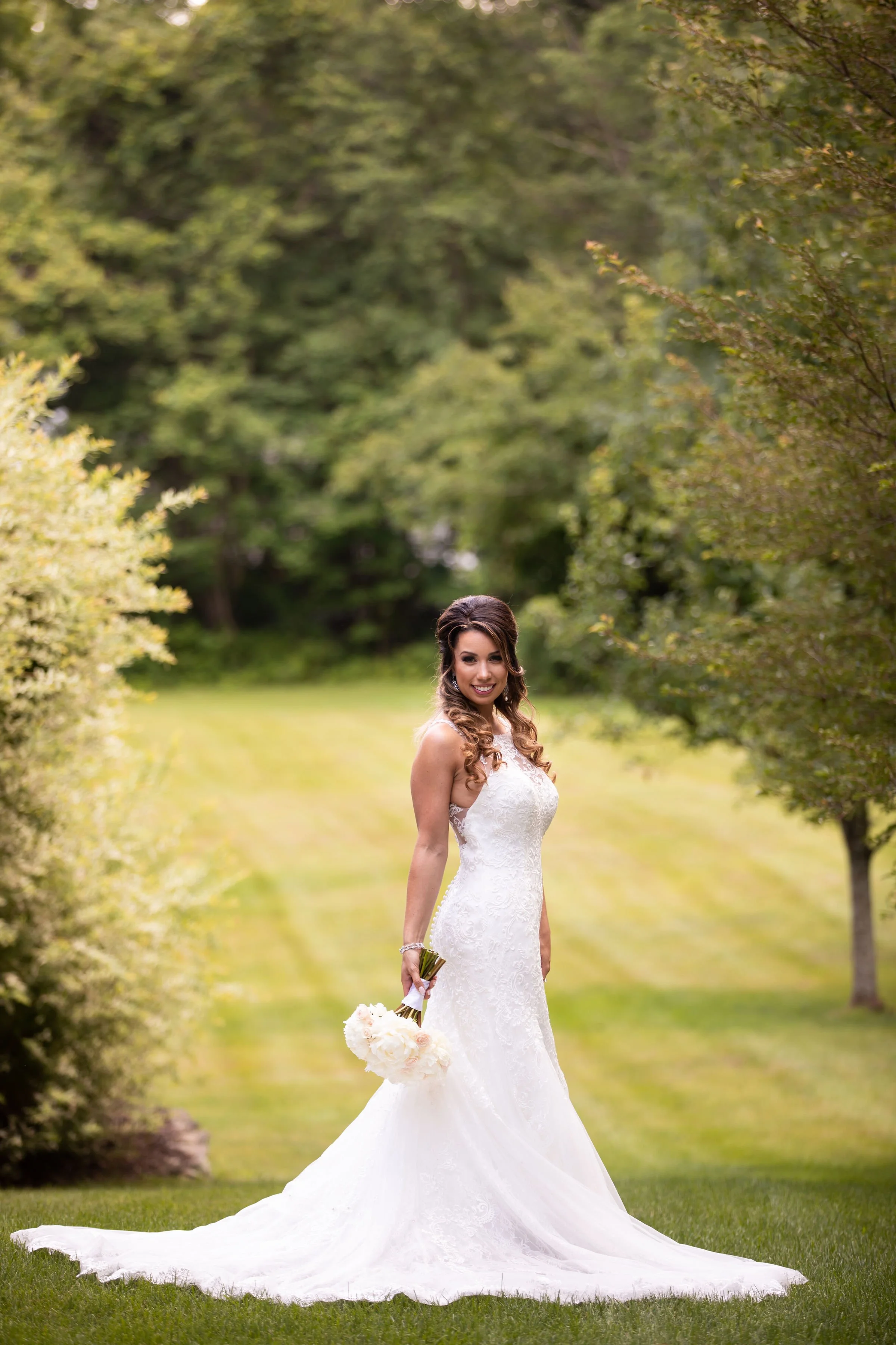 Bride in a white lace wedding dress holding a bouquet of flowers, standing outdoors on a grassy lawn with trees and greenery in the background.