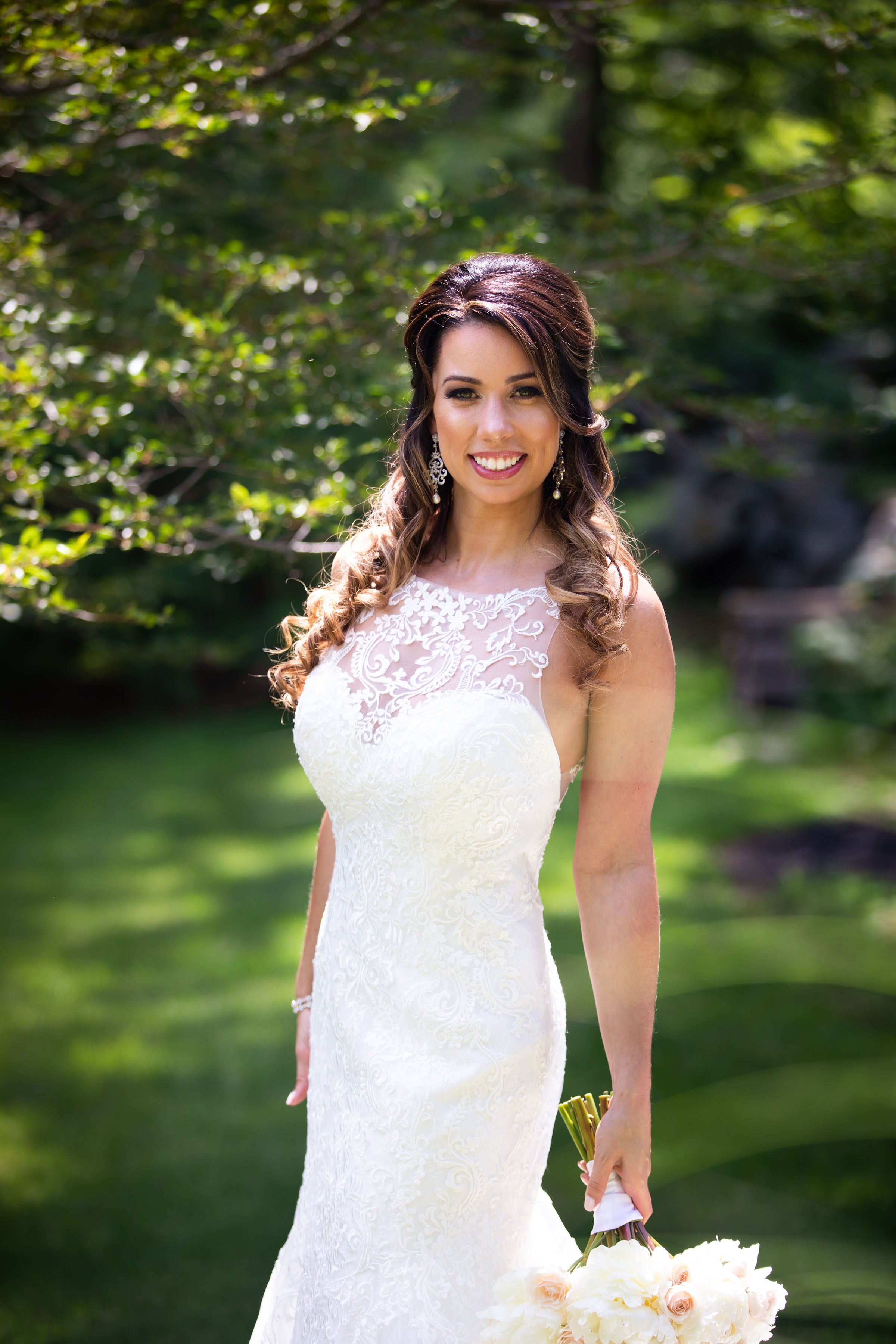 A smiling bride in a white wedding dress holding a bouquet of white and pale pink roses, standing outdoors in a lush green setting with trees and a pond.