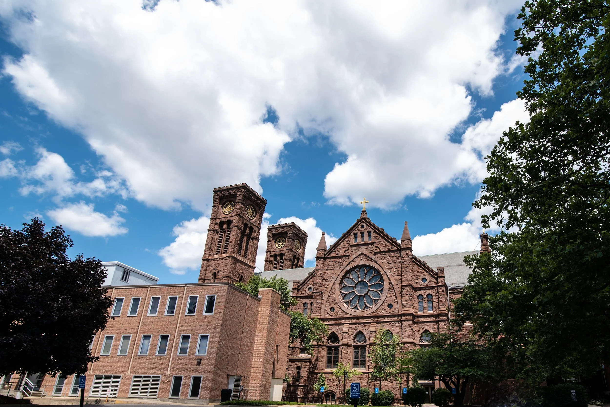 A historic stone church with large circular stained glass window, twin towers with clocks, a cross on top, surrounded by trees, under a partly cloudy sky.