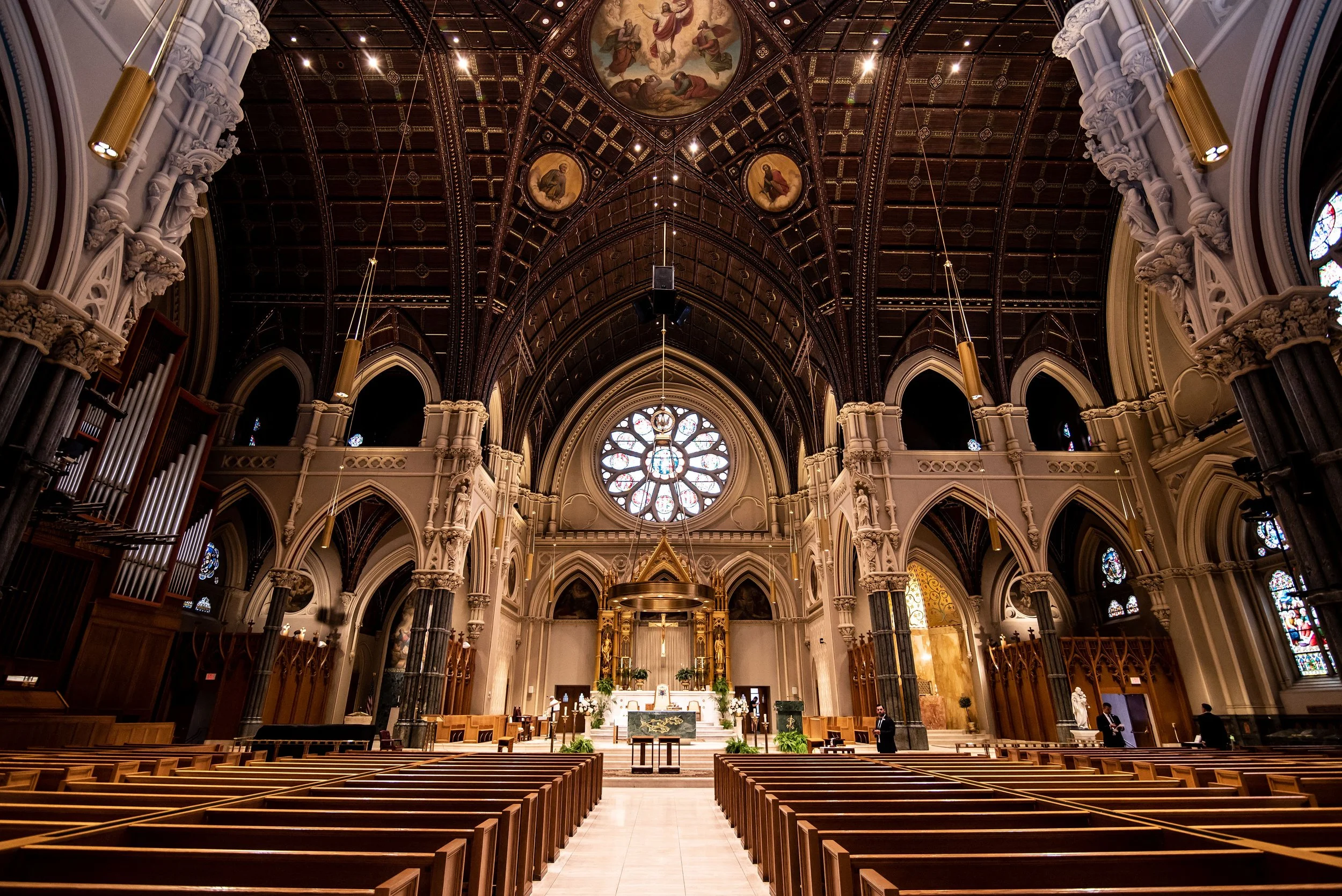 Interior view of a church with detailed architecture, wooden pews, stained glass windows, a large rose window at the front, and an ornate altar.