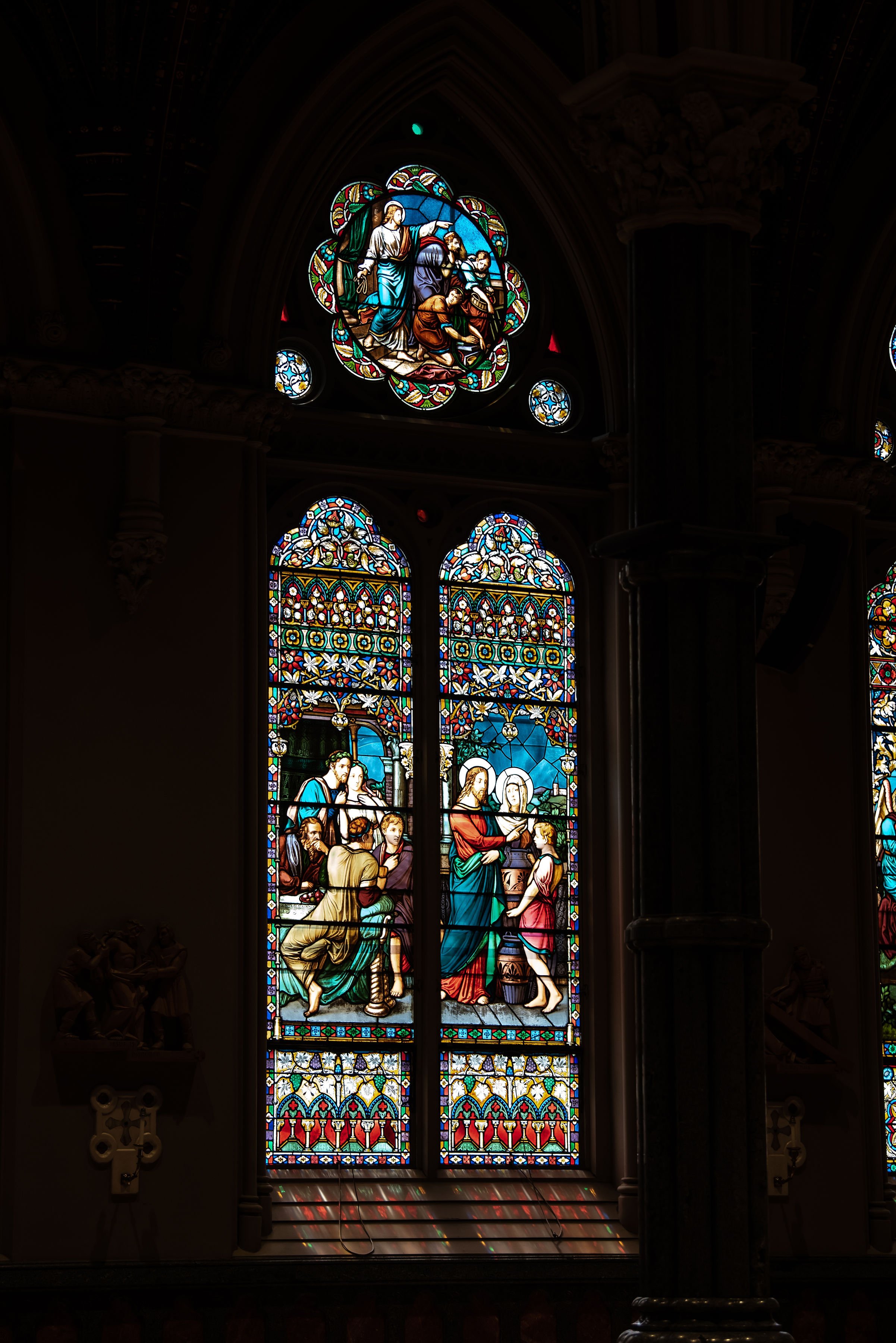 Colorful stained glass windows depicting religious scenes inside a church, with a prominent scene of Jesus with children and a smaller scene above with figures in a biblical setting.