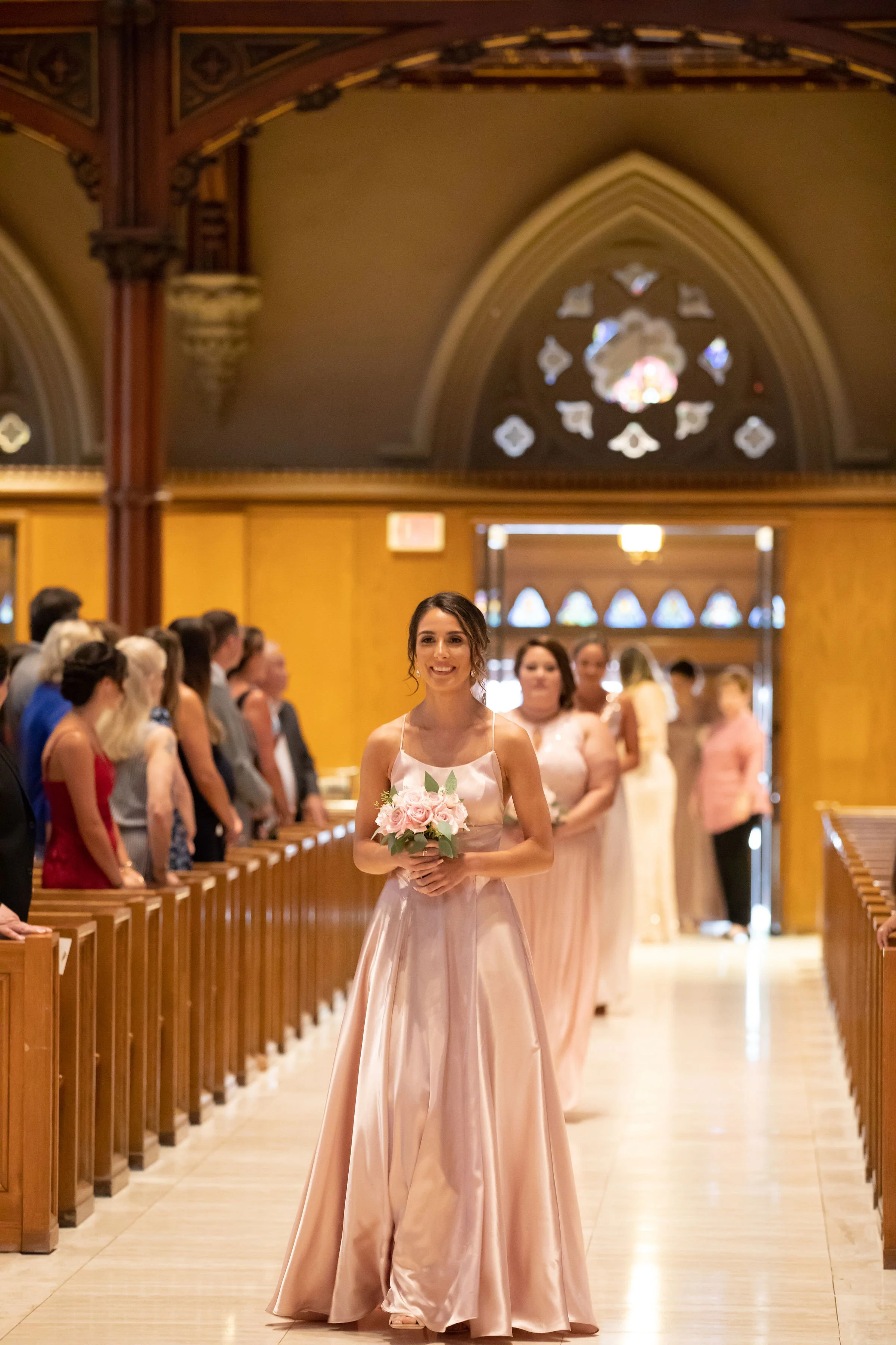 A bride walking down the aisle in a church, holding a bouquet of pink roses, with guests and bridesmaids in the background.