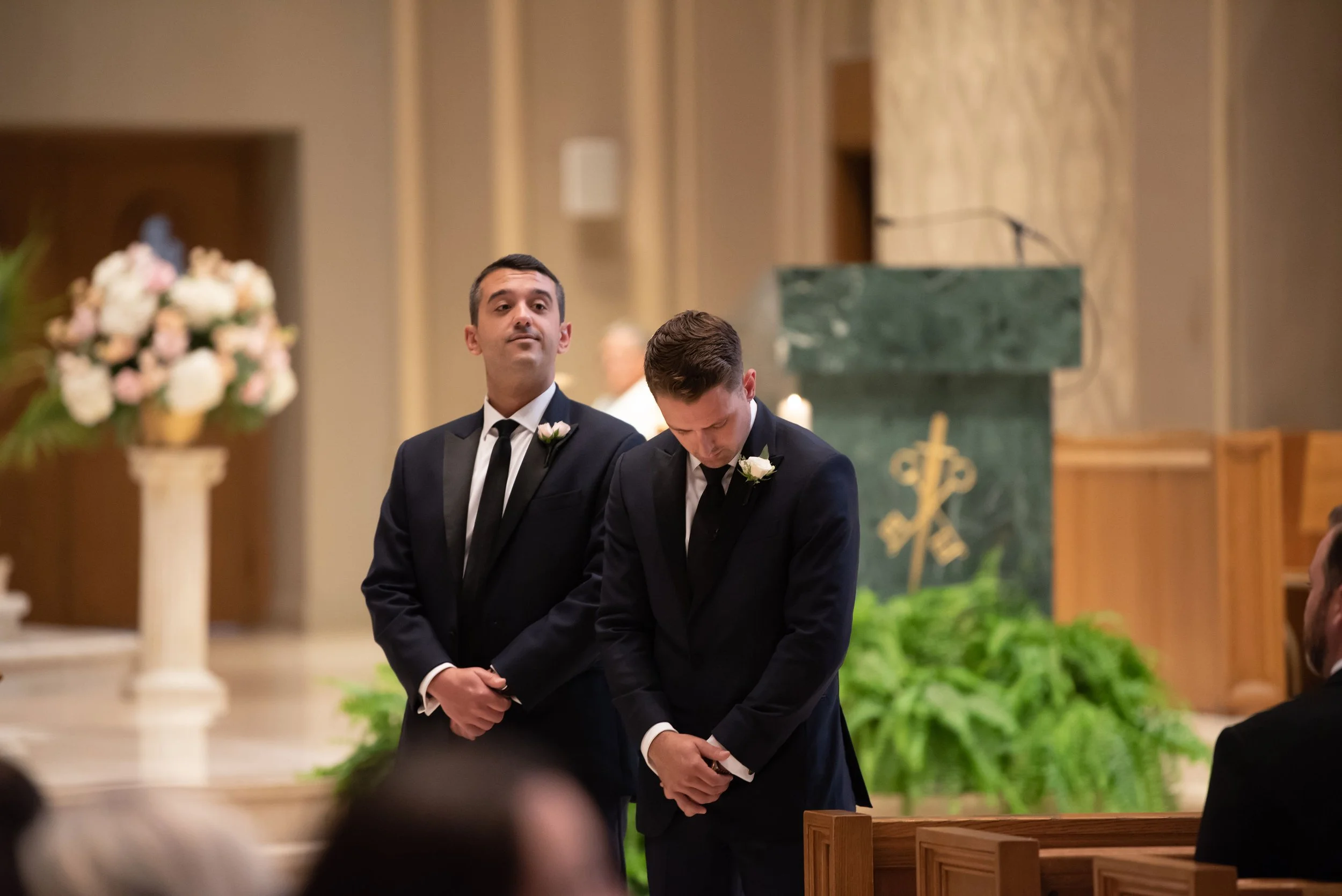 Two men in black suits and white shirts at a wedding ceremony, one standing with hands clasped and the other bowing his head in prayer or reflection, inside a church with floral arrangements and a green marble altar in the background.