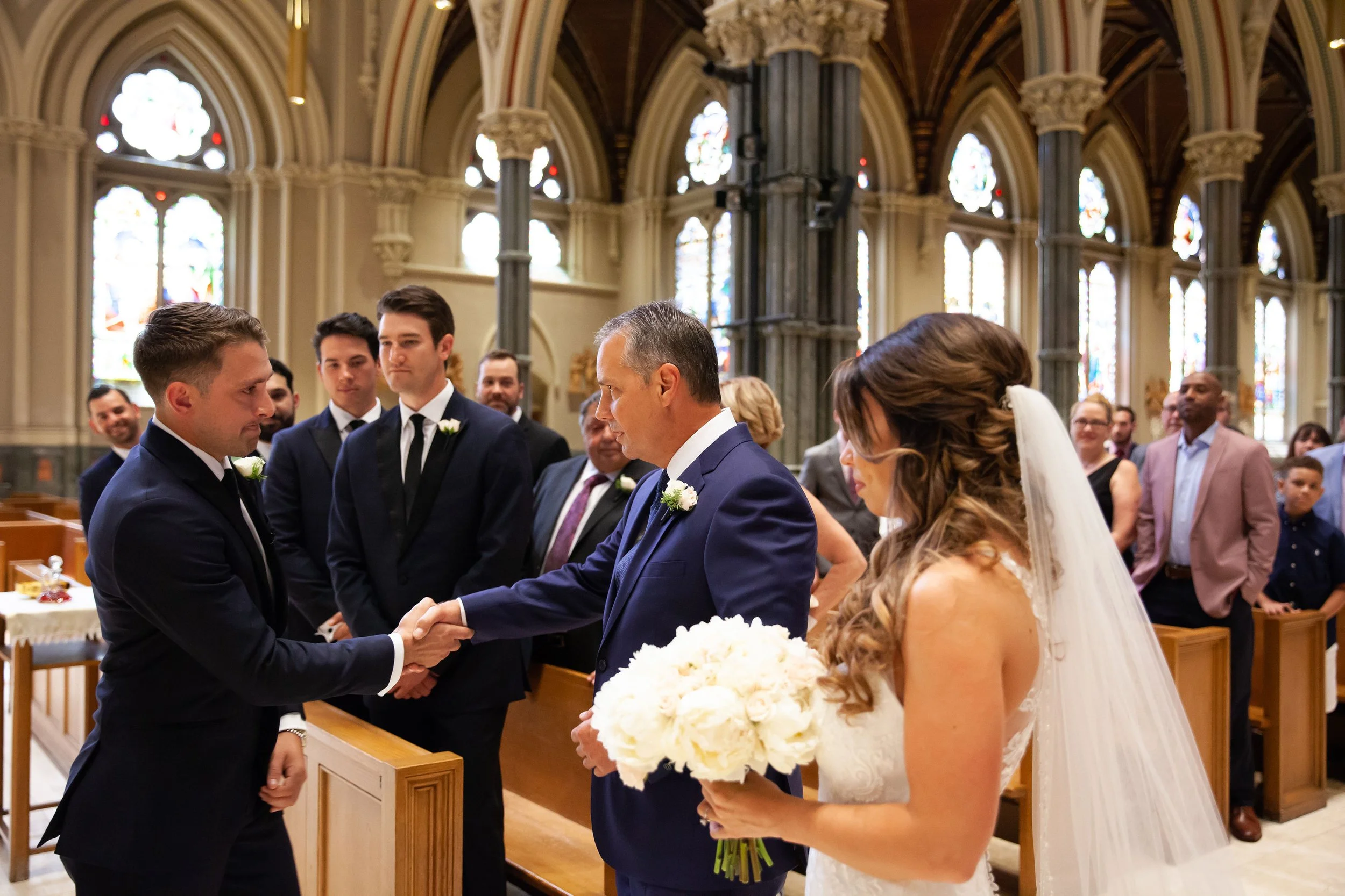 Bride and groom shaking hands with a priest during wedding ceremony in a church, with guests observing.