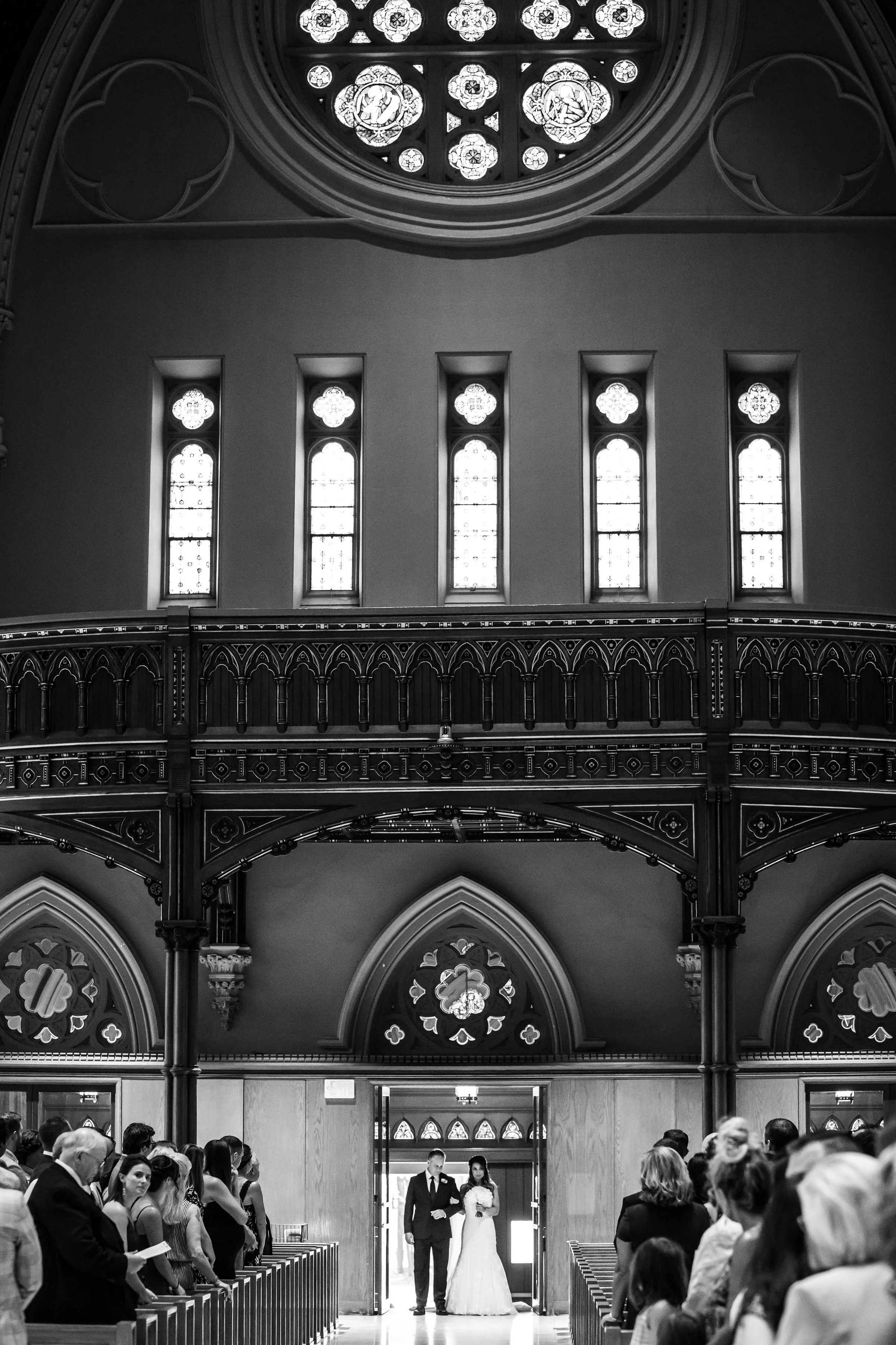 Black and white photo of a wedding ceremony inside a church, with a bride and groom standing at the entrance, guests seated on either side, and ornate stained glass windows above.