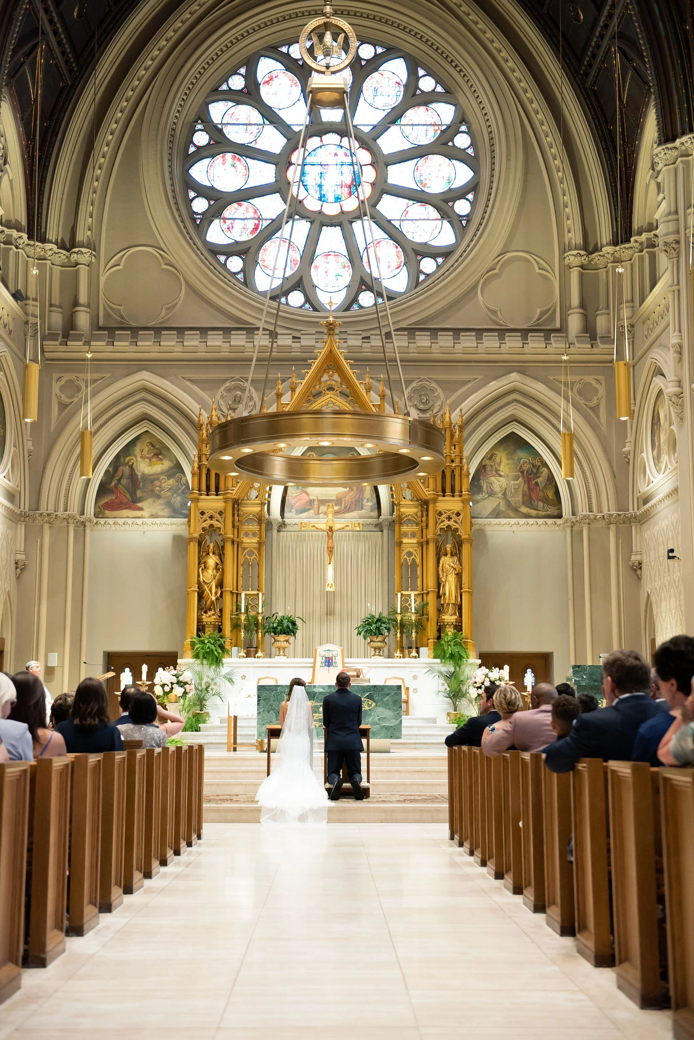 A wedding ceremony taking place inside a grand church, with a bride and groom kneeling at the altar facing the priest. The church has a large circular stained glass window, ornate gold accents, religious statues, and decorated walls.