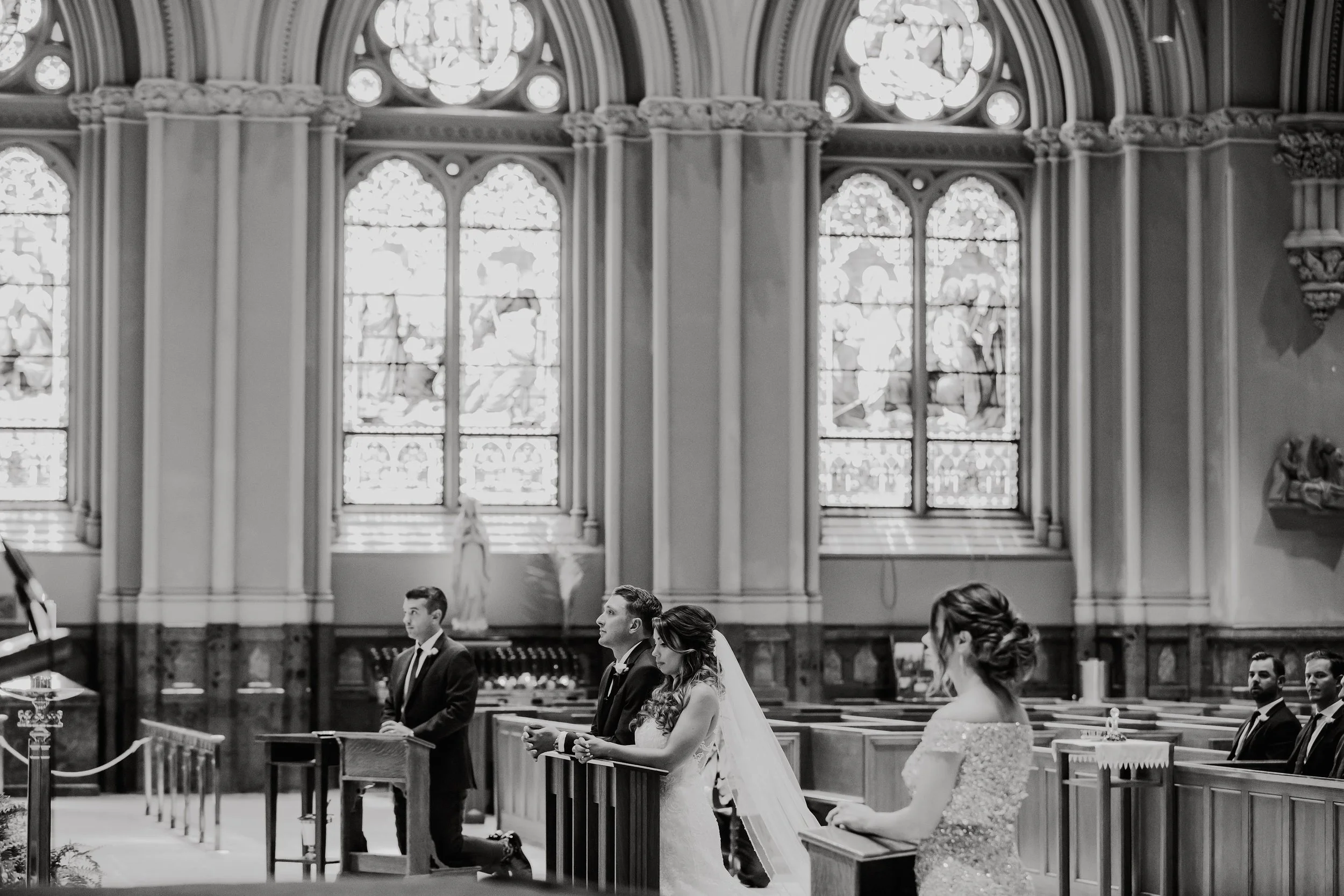 Black and white photo of a wedding ceremony inside a church, with a bride, groom, and attendants standing at the altar in front of stained glass windows.