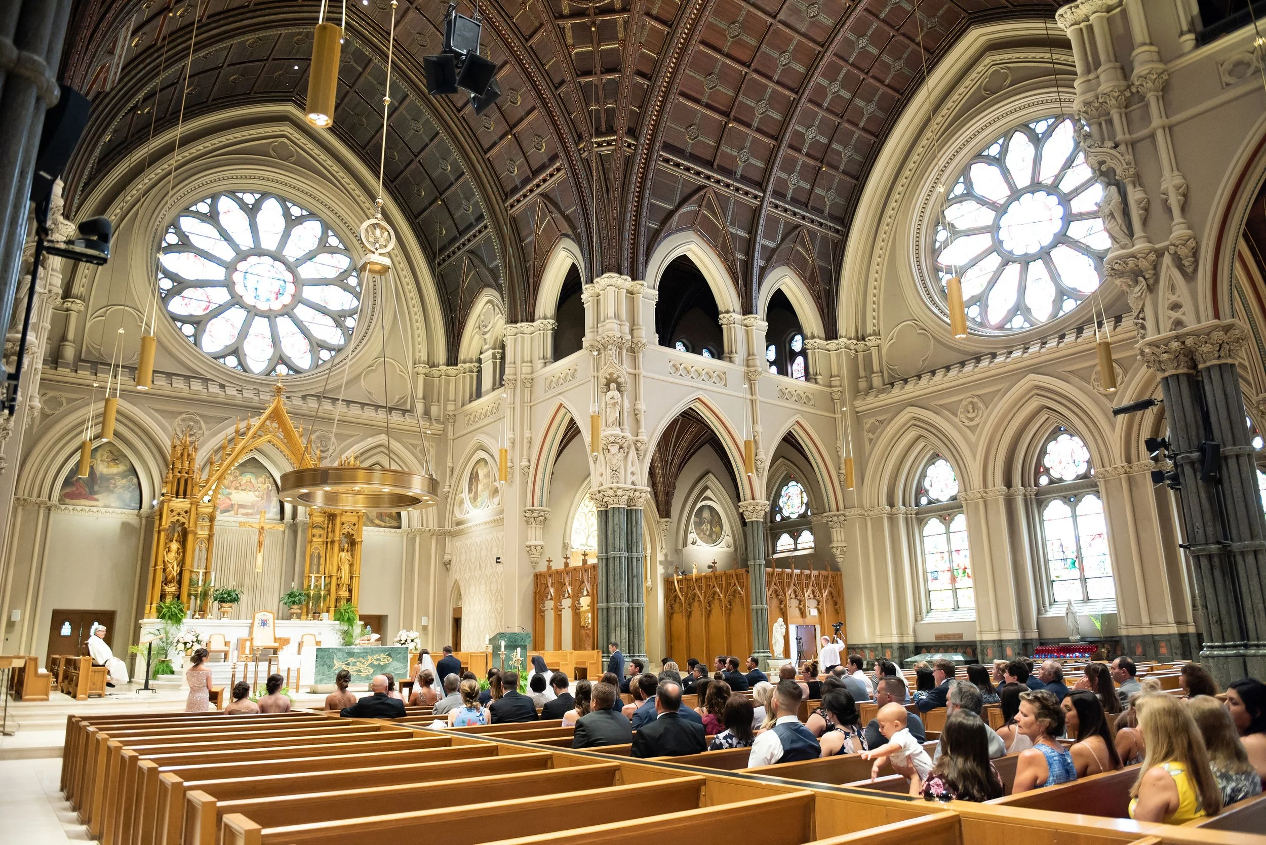 Interior of a church during a wedding service, with guests seated in pews, altar at front, stained glass windows, and ornate architecture.