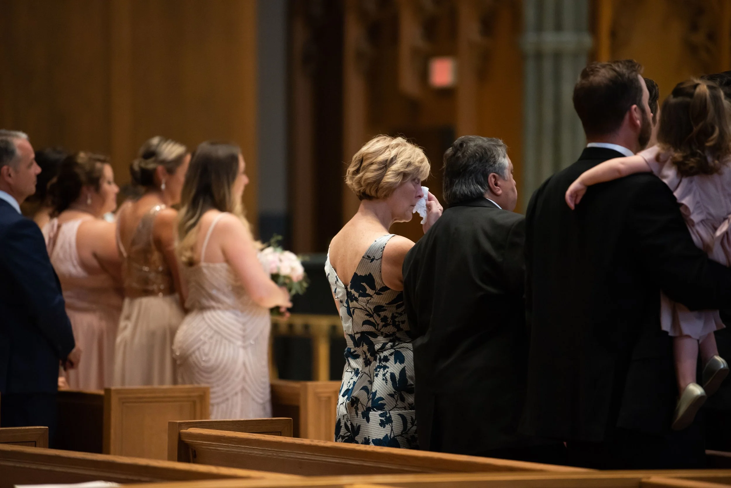 People standing inside a church during a wedding ceremony, some holding flowers, with a woman wiping tears from her eyes.