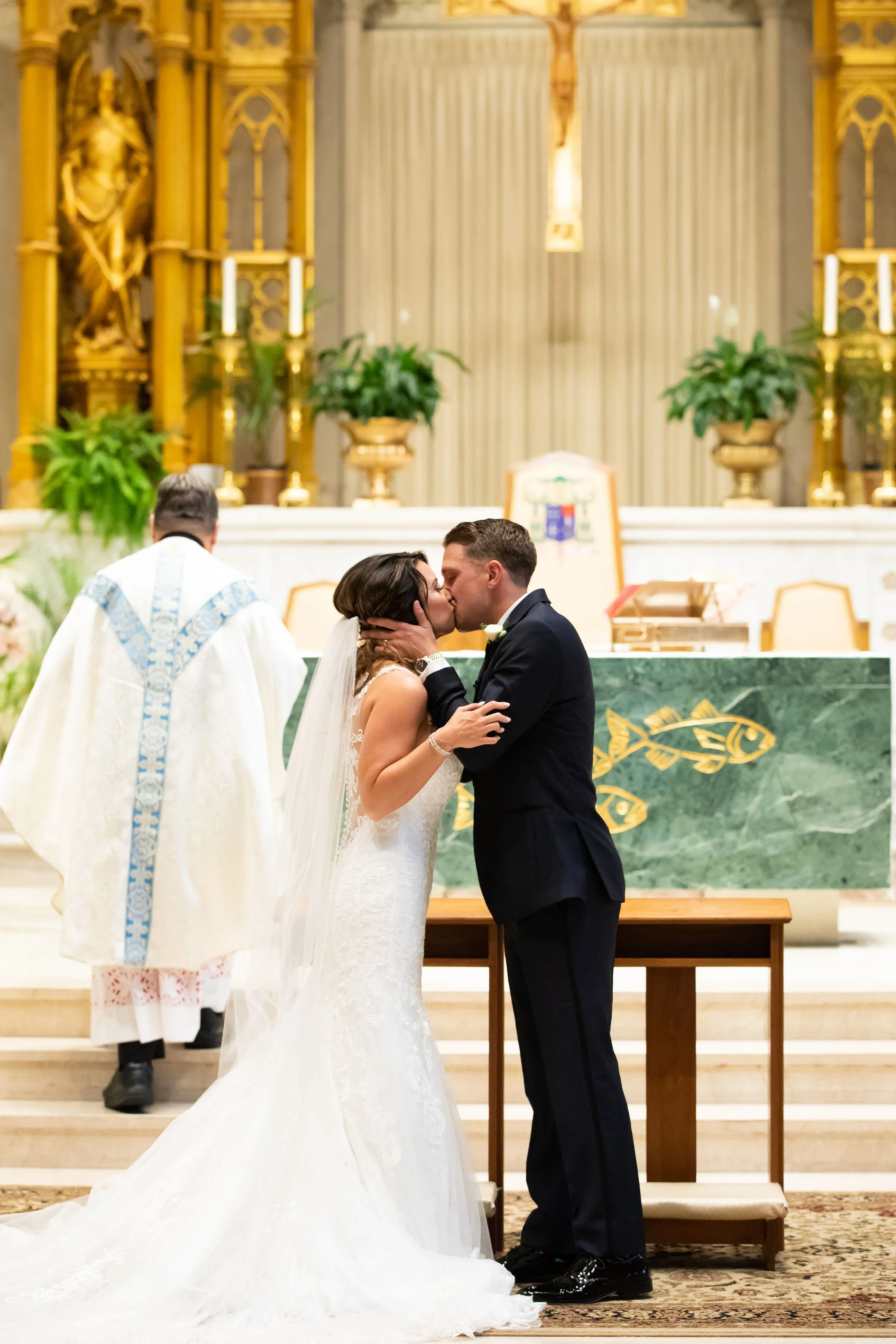 A bride and groom exchange a kiss during their wedding ceremony inside a church. The bride wears a white wedding gown and veil, and the groom wears a black tuxedo. A priest stands behind them, officiating the ceremony, with church altar and decoratio
