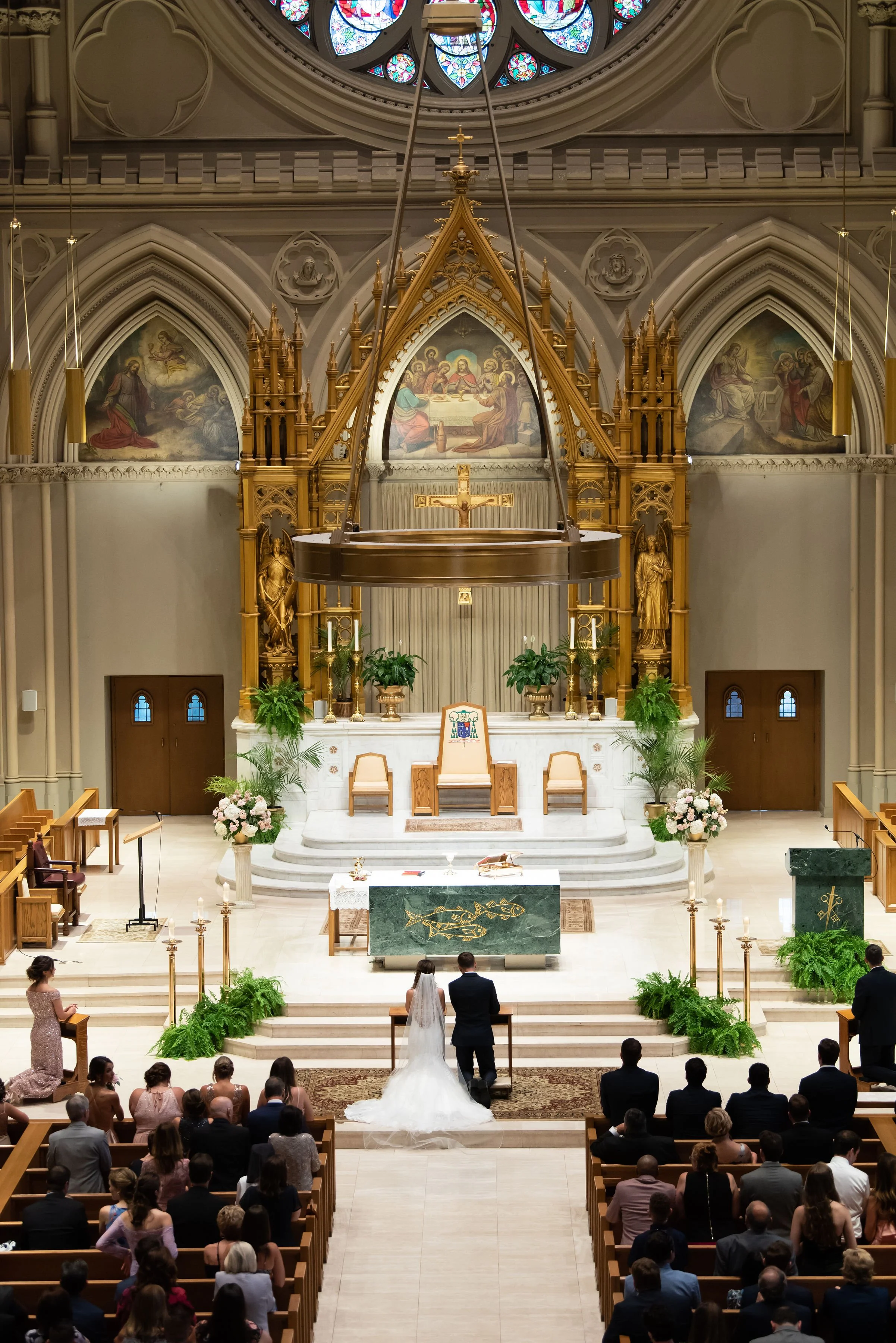 A wedding ceremony taking place inside a church with a bride and groom standing at the altar, surrounded by guests seated in pews, with religious artwork and ornate decorations in the background.