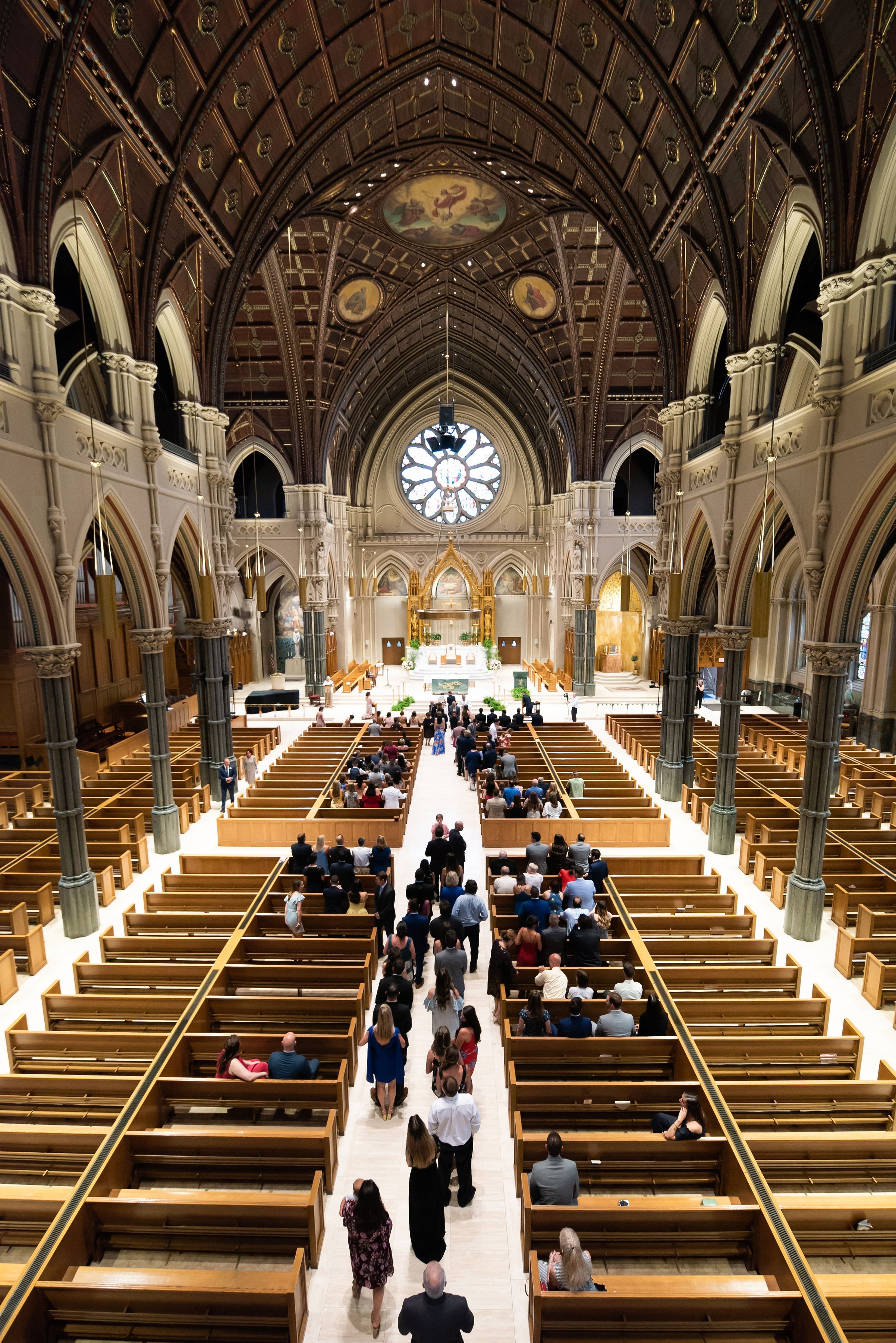 Interior view of a church filled with people sitting on wooden pews, with a focus on the altar at the front, featuring stained glass windows and ornate architecture.
