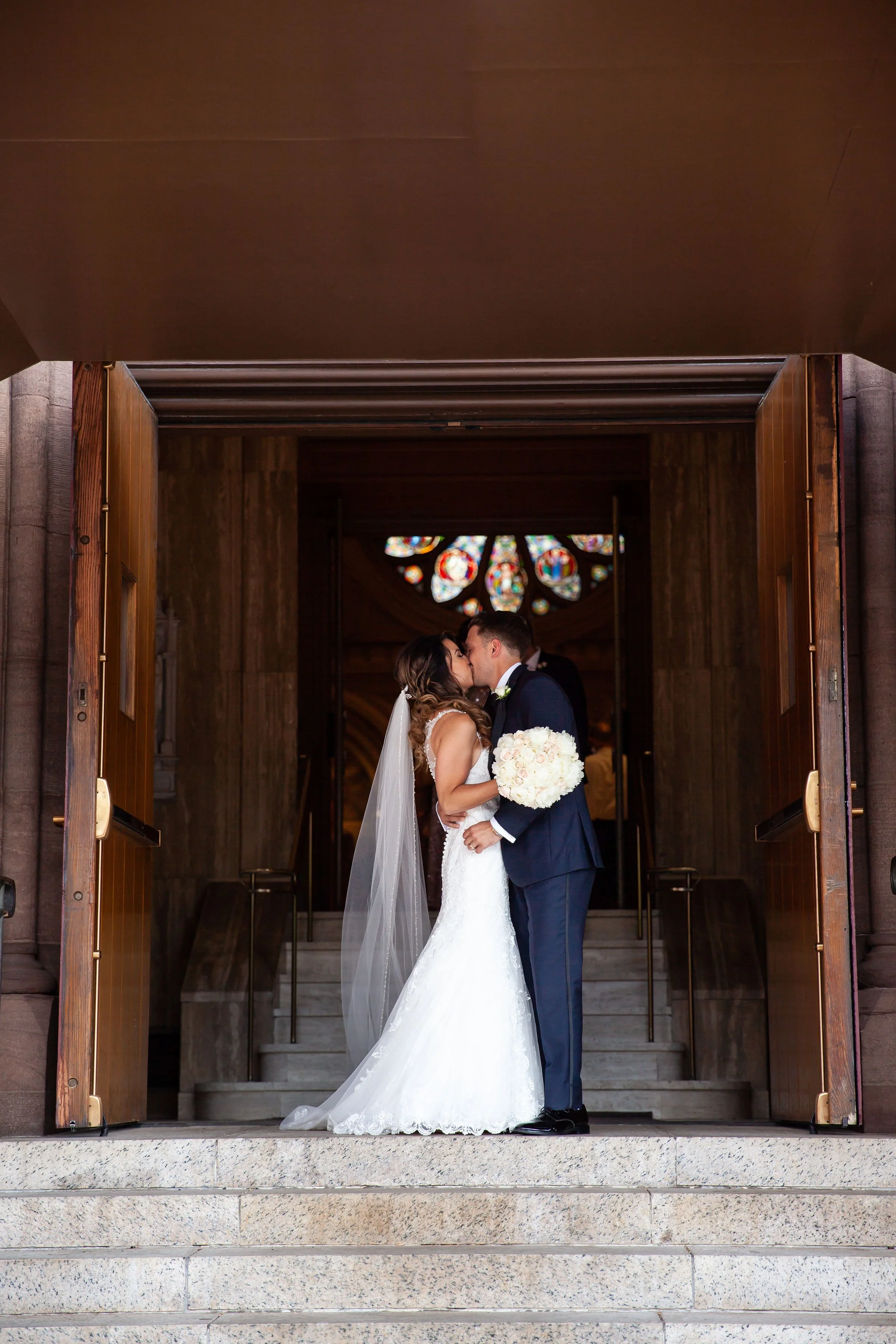 A newlywed couple sharing a kiss in front of church doors with a stained glass window in the background.