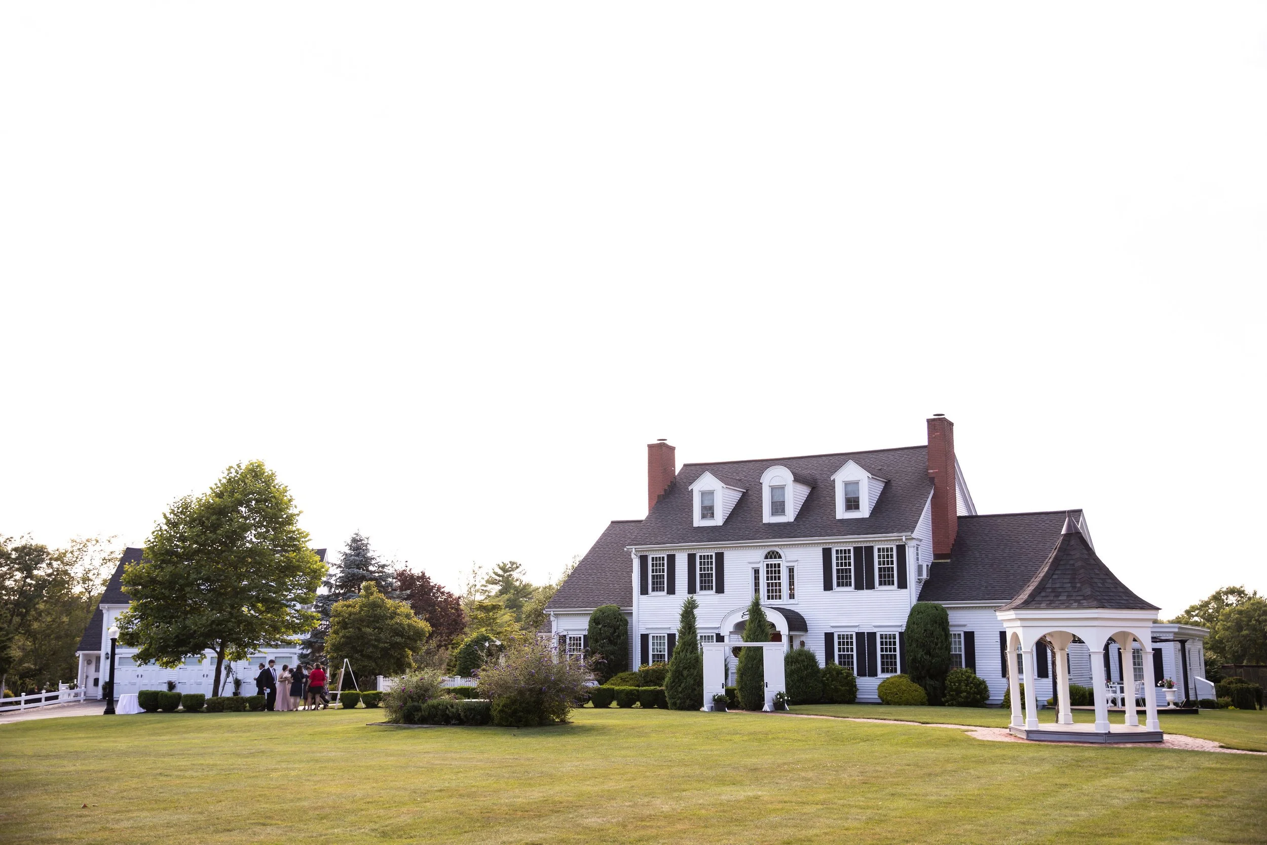 Large white historic house with black shutters, a turret, and a garden, possibly for a wedding or event, with a group of people gathered near the house and trees in the background.