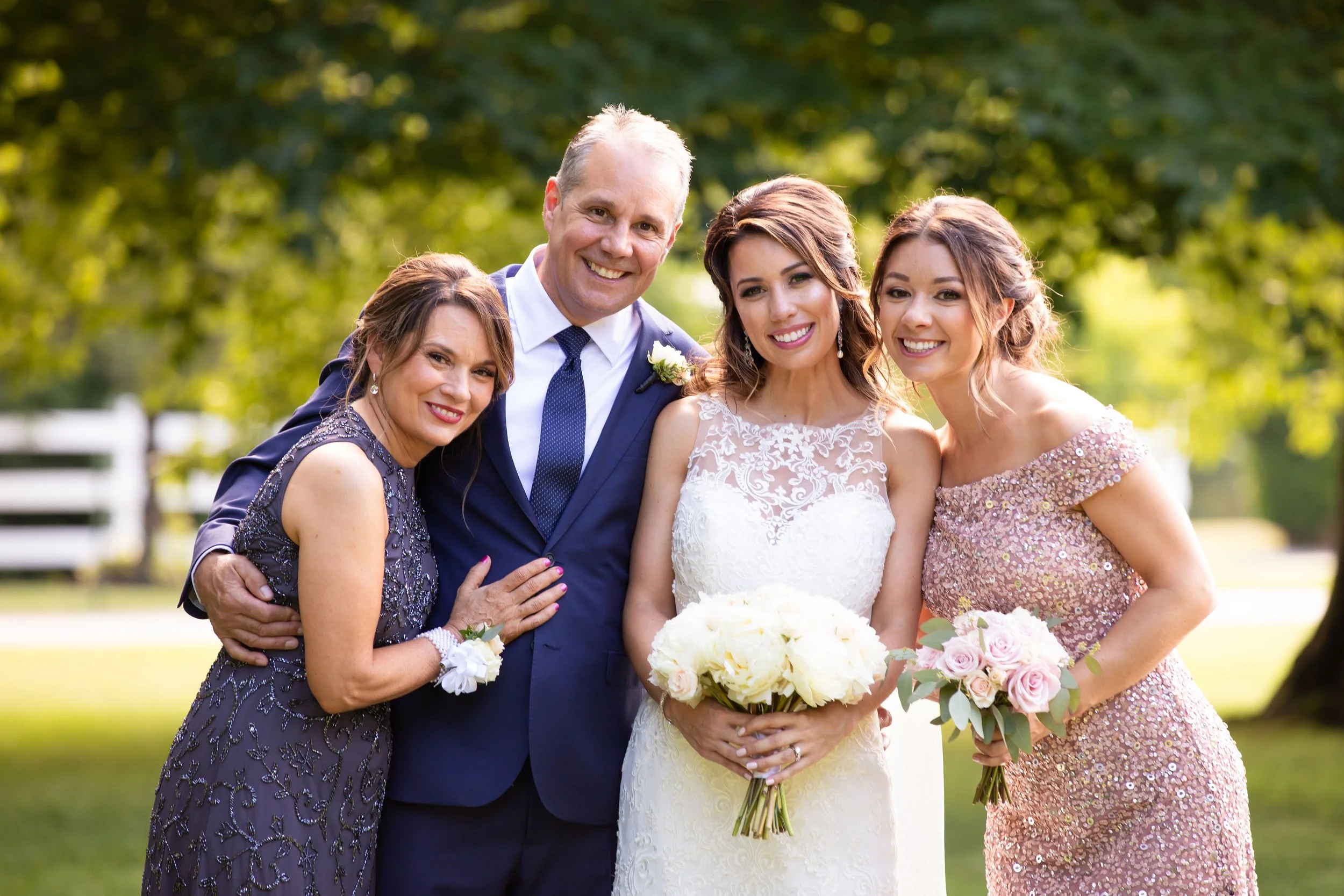 A group of four people, two women and two men, at a wedding outdoors. They are smiling and dressed in formal attire, with the bride holding a bouquet of white peonies and the other woman holding pink roses. The setting features green trees and a brig
