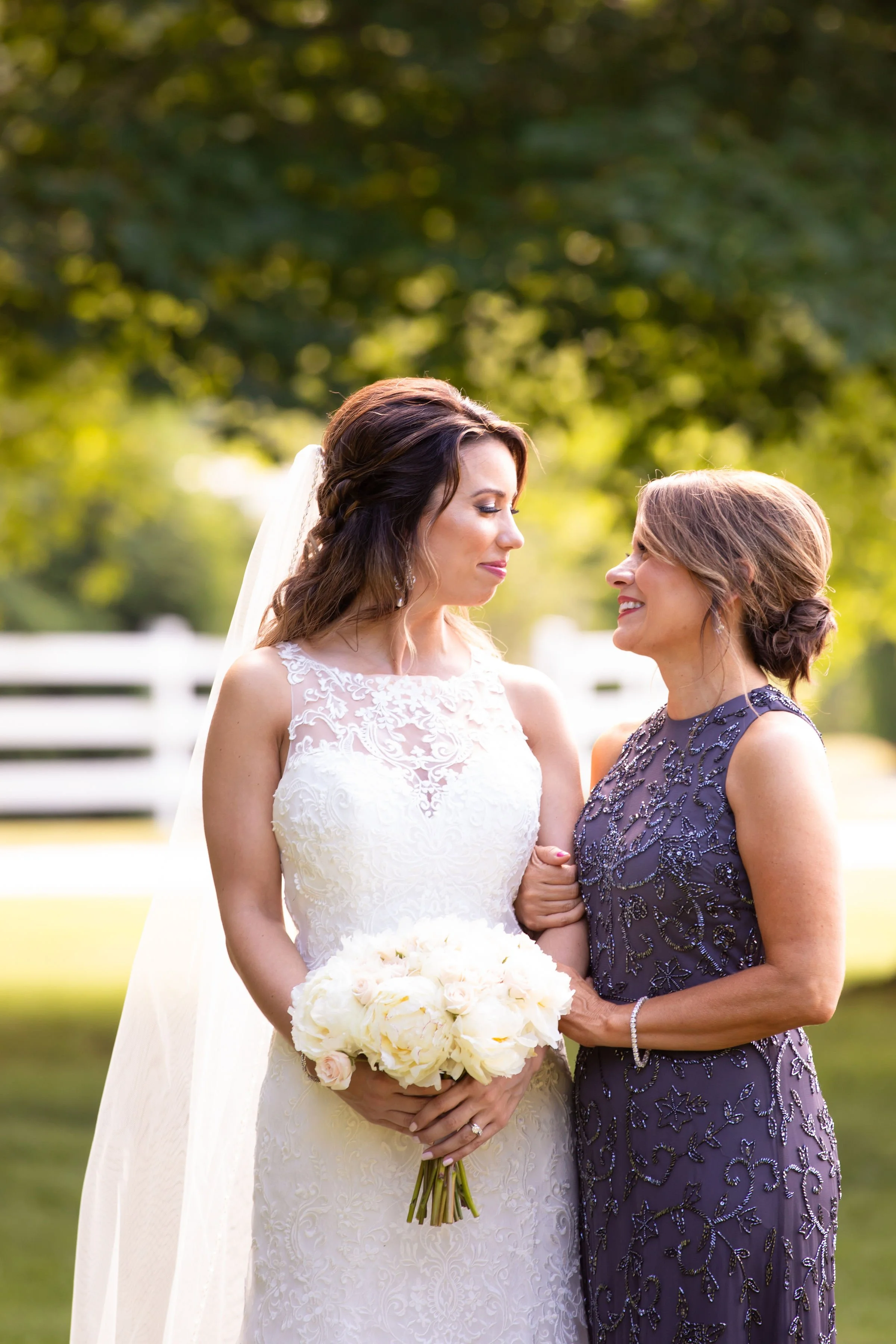 A bride in a white lace wedding dress holding a bouquet of white flowers, standing next to a woman in a dark purple embroidered dress, with their faces close together and smiling outdoors during a sunny day.