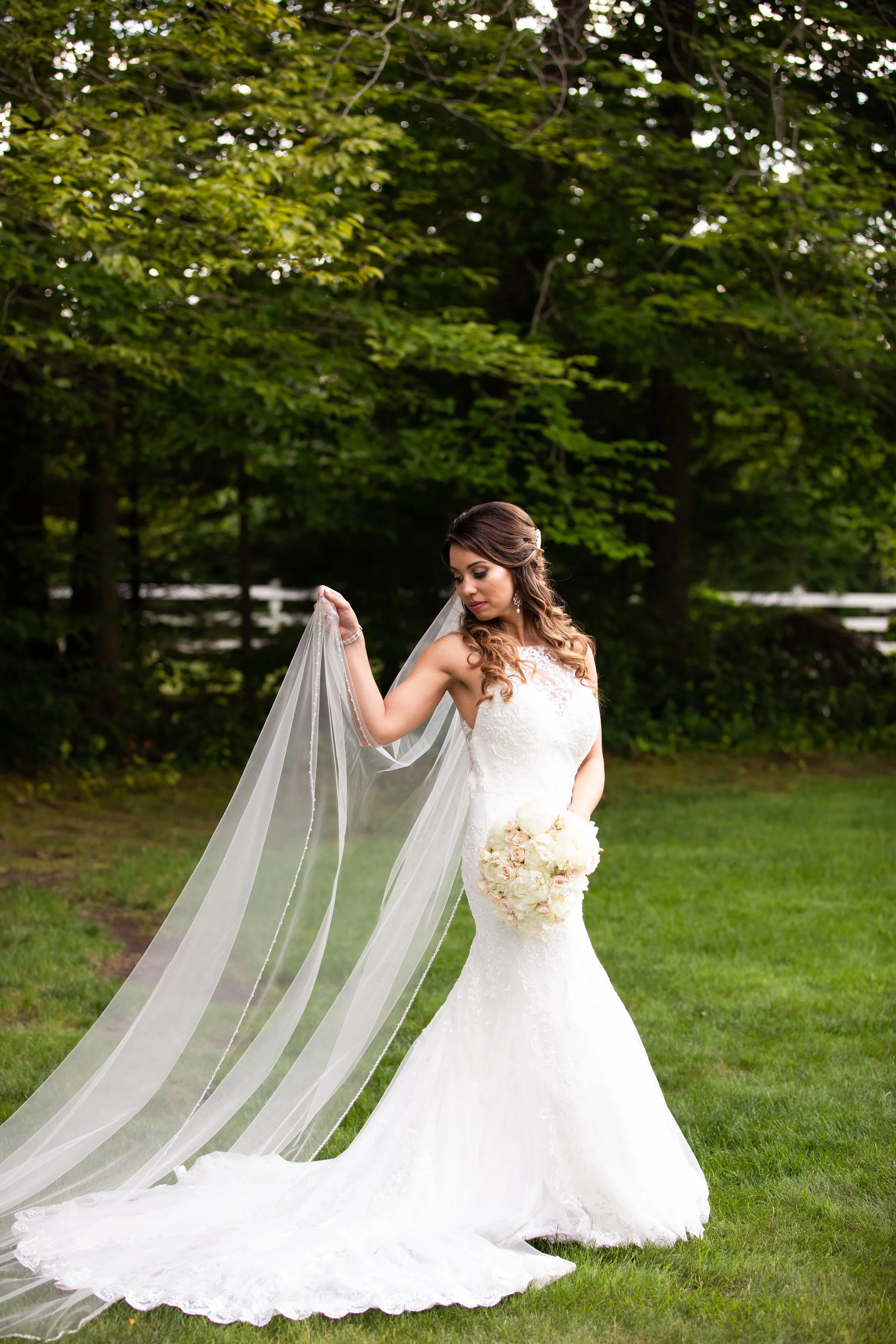 A bride in a white wedding gown holding a bouquet of white and pale pink flowers outdoors with greenery in the background.