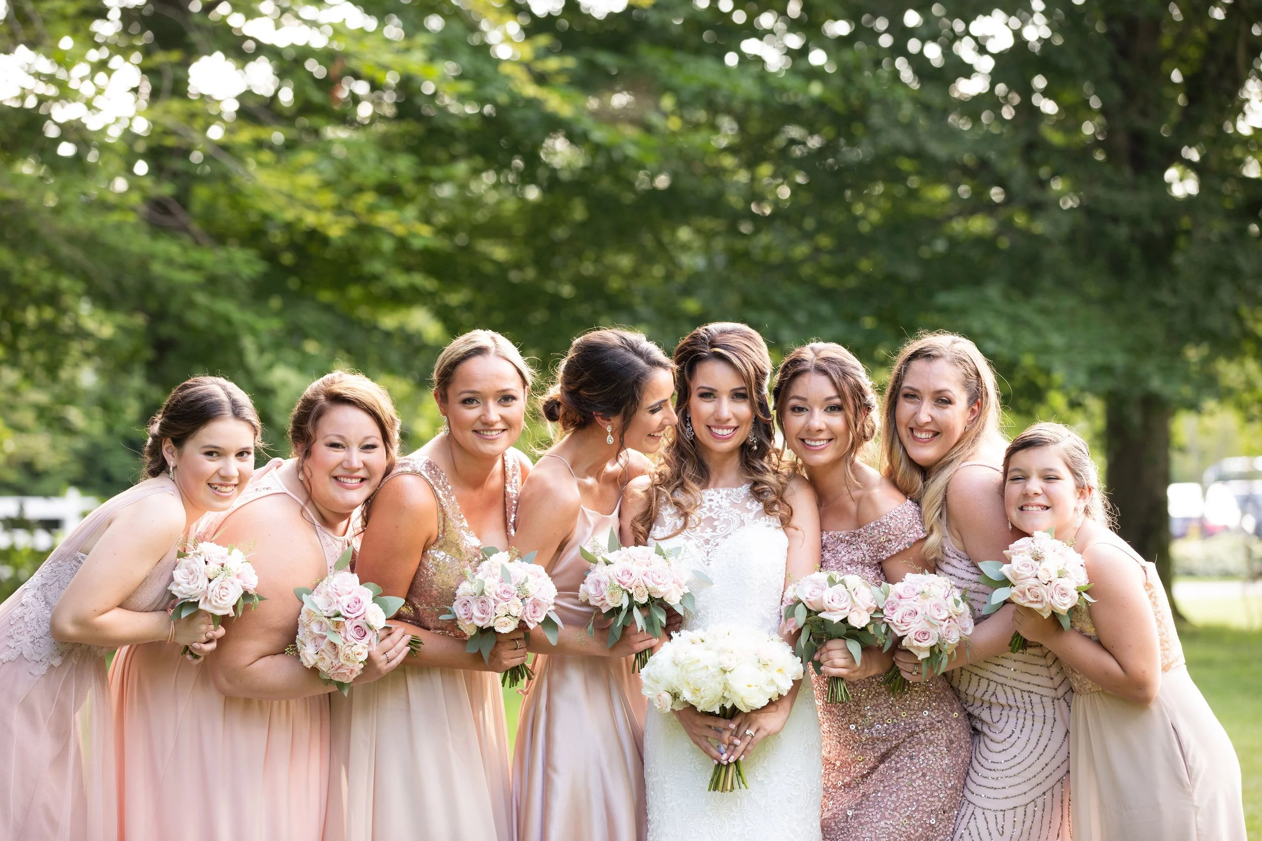 A bride and her bridesmaids posing outdoors, holding bouquets, on a sunny day with green trees in the background.