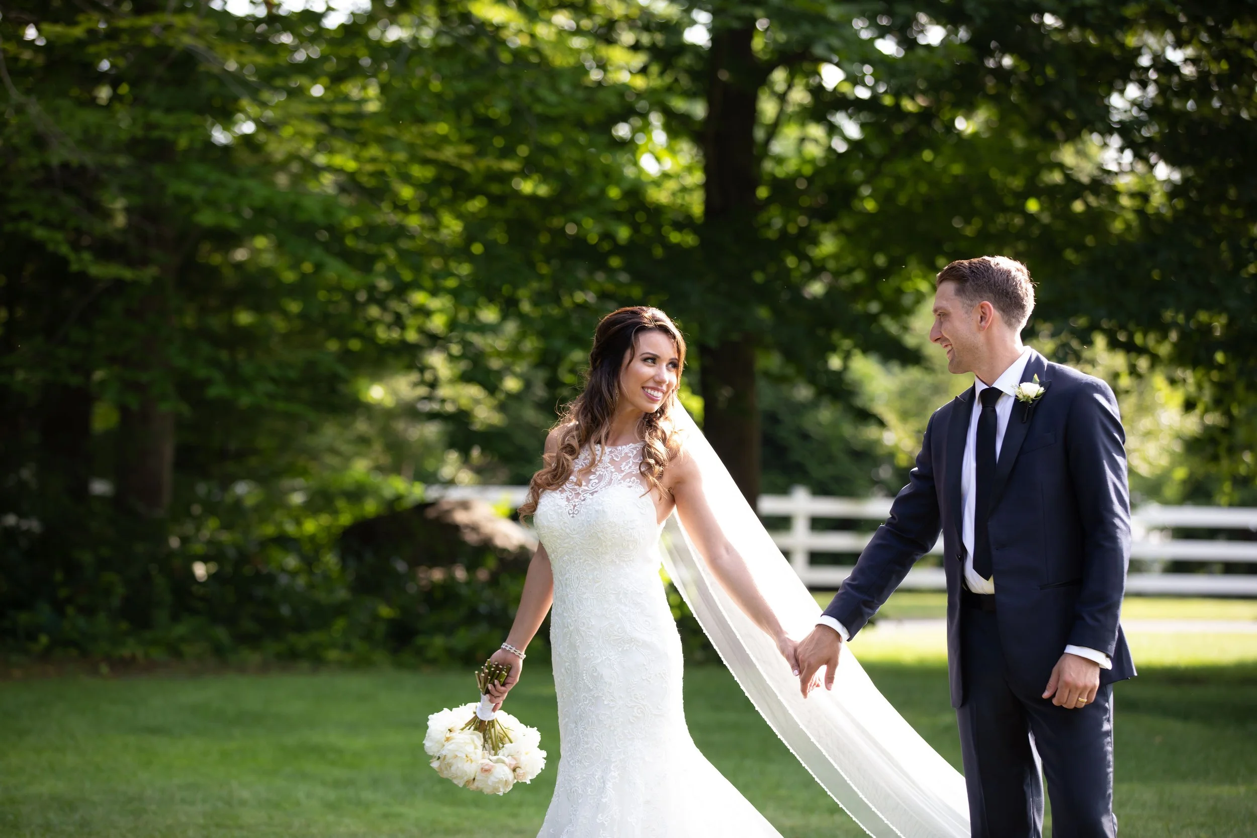 A bride and groom holding hands outdoors on a lush green lawn with trees and a white fence in the background, smiling at each other.