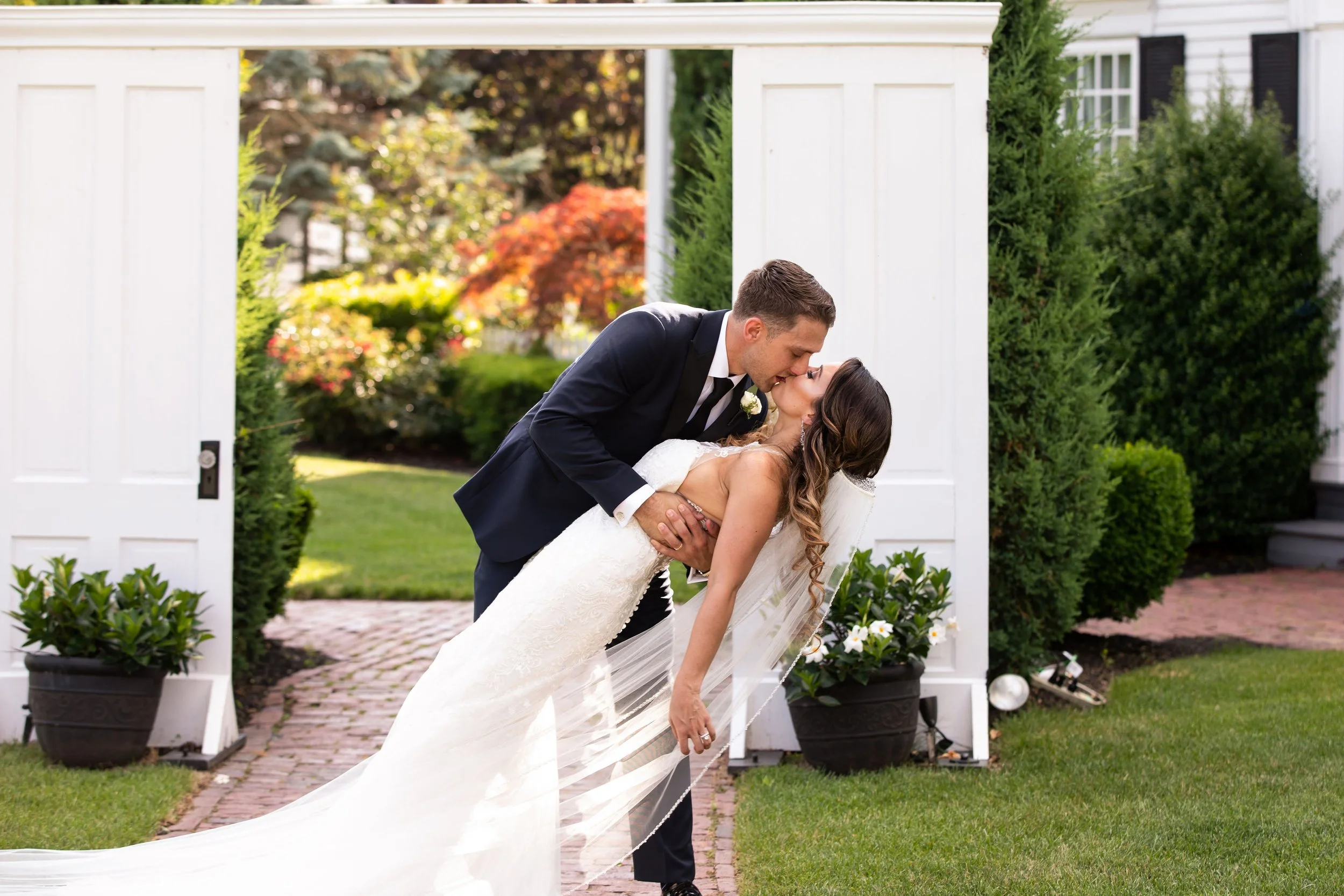A newlywed couple shares a kiss outdoors in front of a white wooden gate, surrounded by lush green plants and colorful autumn trees.