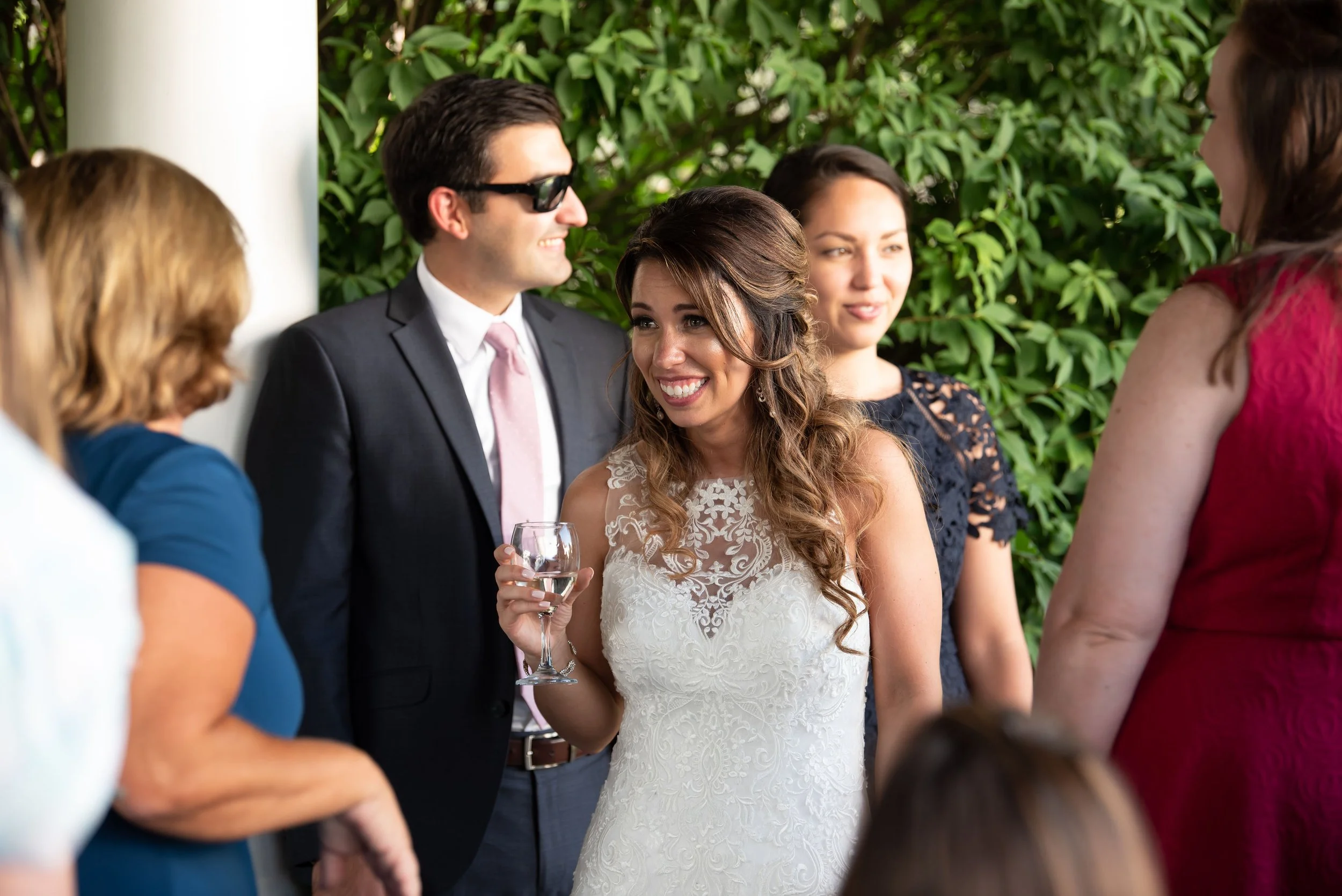 A bride holding a wine glass smiling and talking to a group of people at her wedding reception, with greenery in the background.