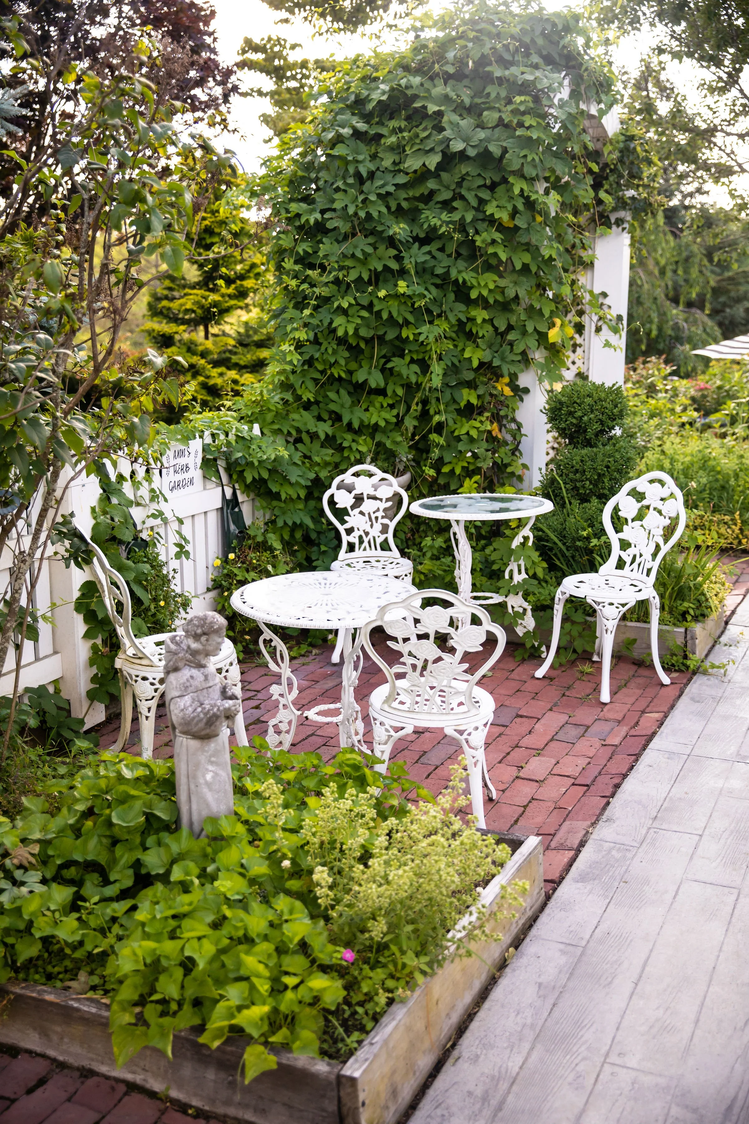 An outdoor garden scene with white wrought iron patio furniture, including a small round table and four chairs, situated on a brick patio. The surrounding lush greenery includes a hedge, a potted plant, and various flowering plants. There is a small 