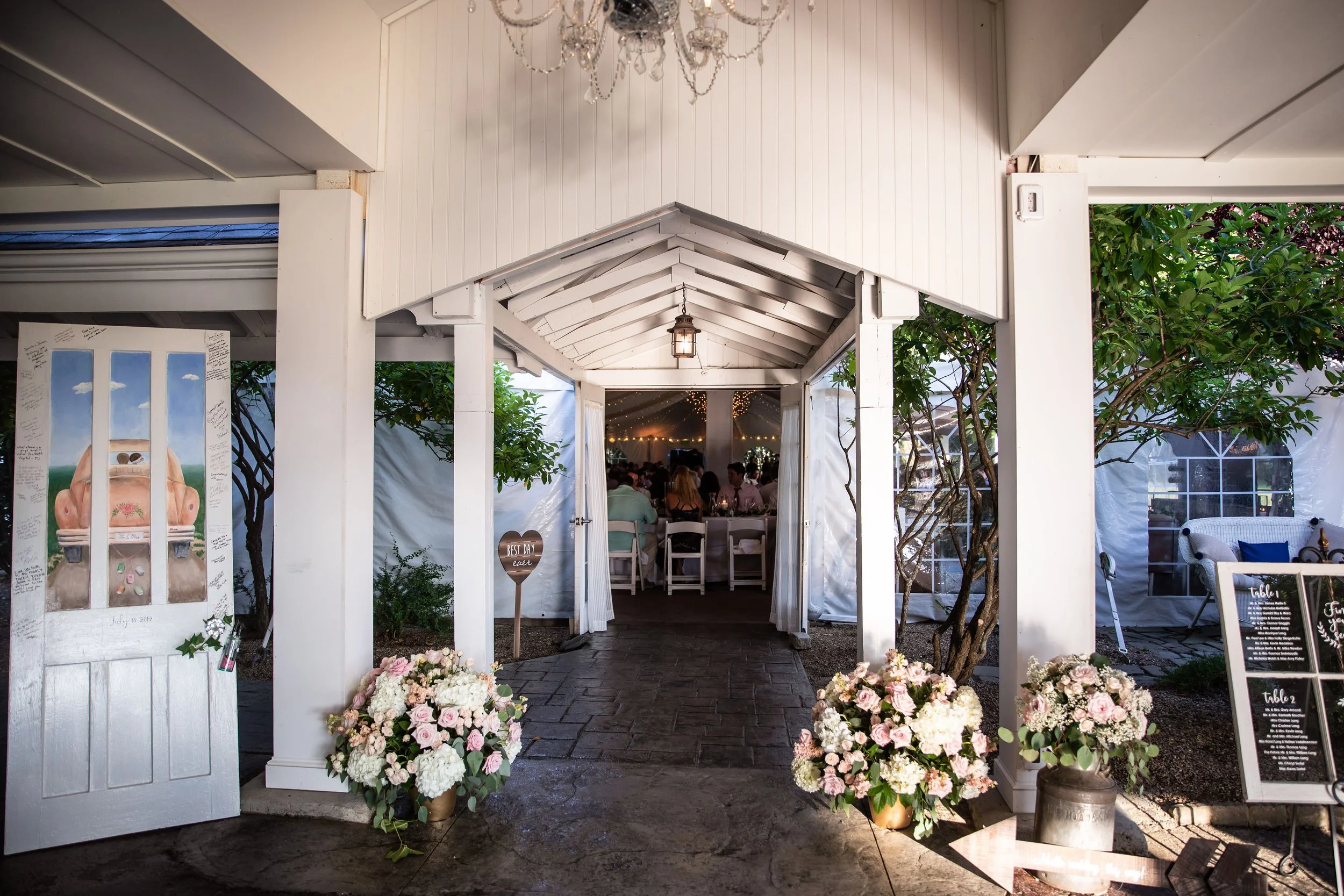 Entrance to a wedding reception with white wooden frame, floral arrangements, and a view of people sitting at tables inside under string lights.