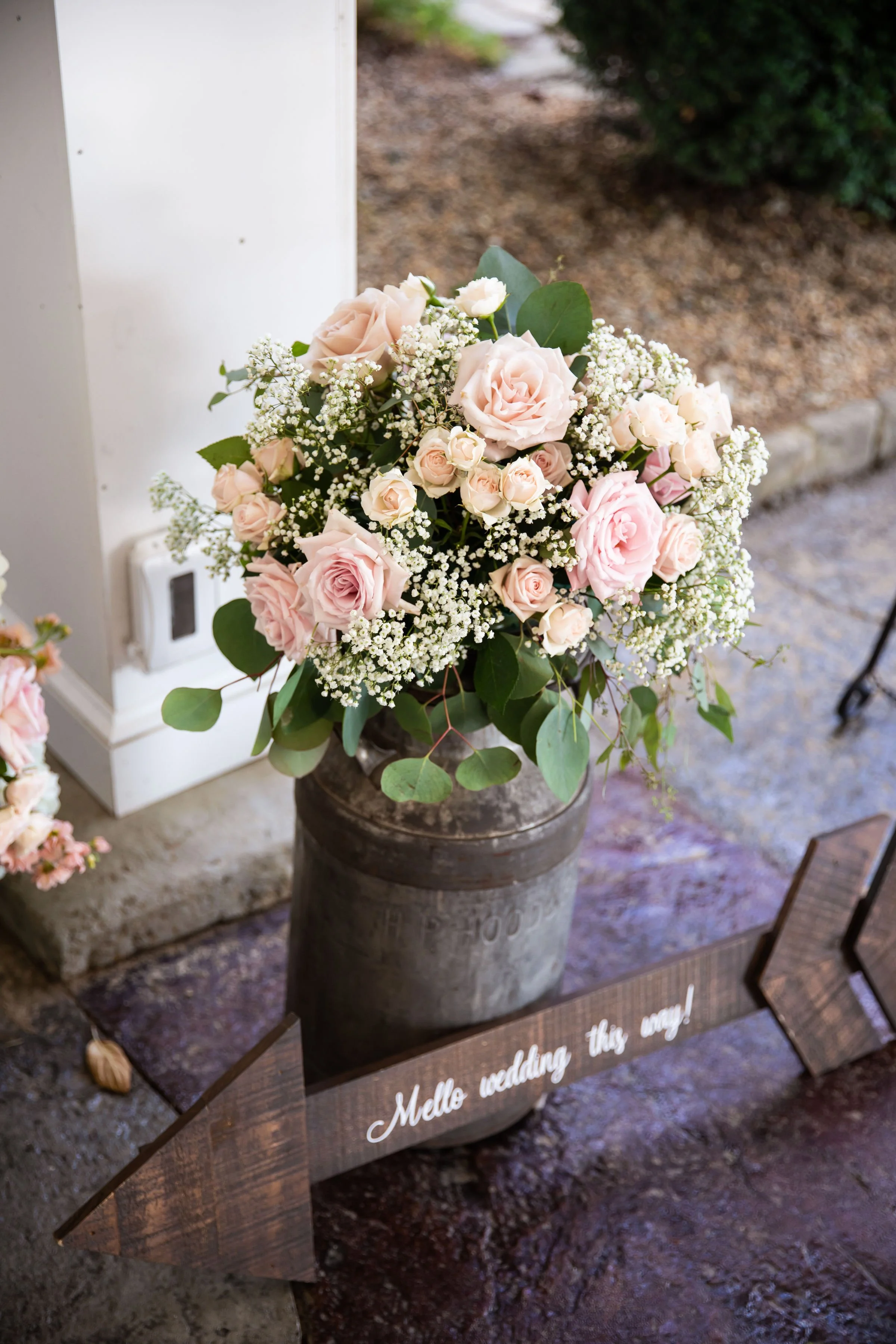 A bouquet of light pink roses and white baby's breath flowers arranged in a dark vase, placed on a vintage metal milk can on a concrete floor. There is a sign in front with the text "Mello wedding this way!"