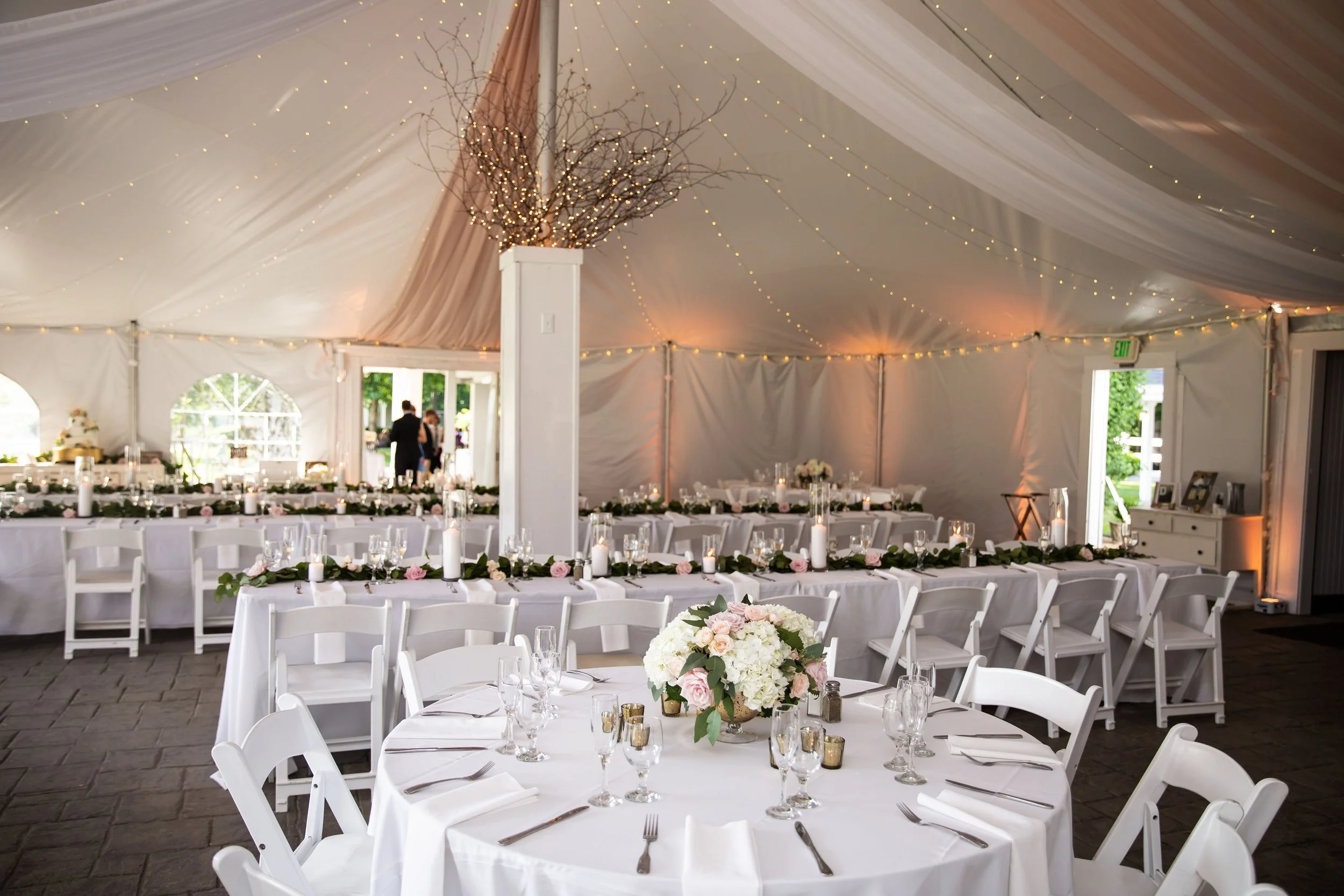 Elegant wedding reception tent with round tables and flower centerpieces, decorated with candles and white chairs, illuminated with string lights and soft pink drapery.