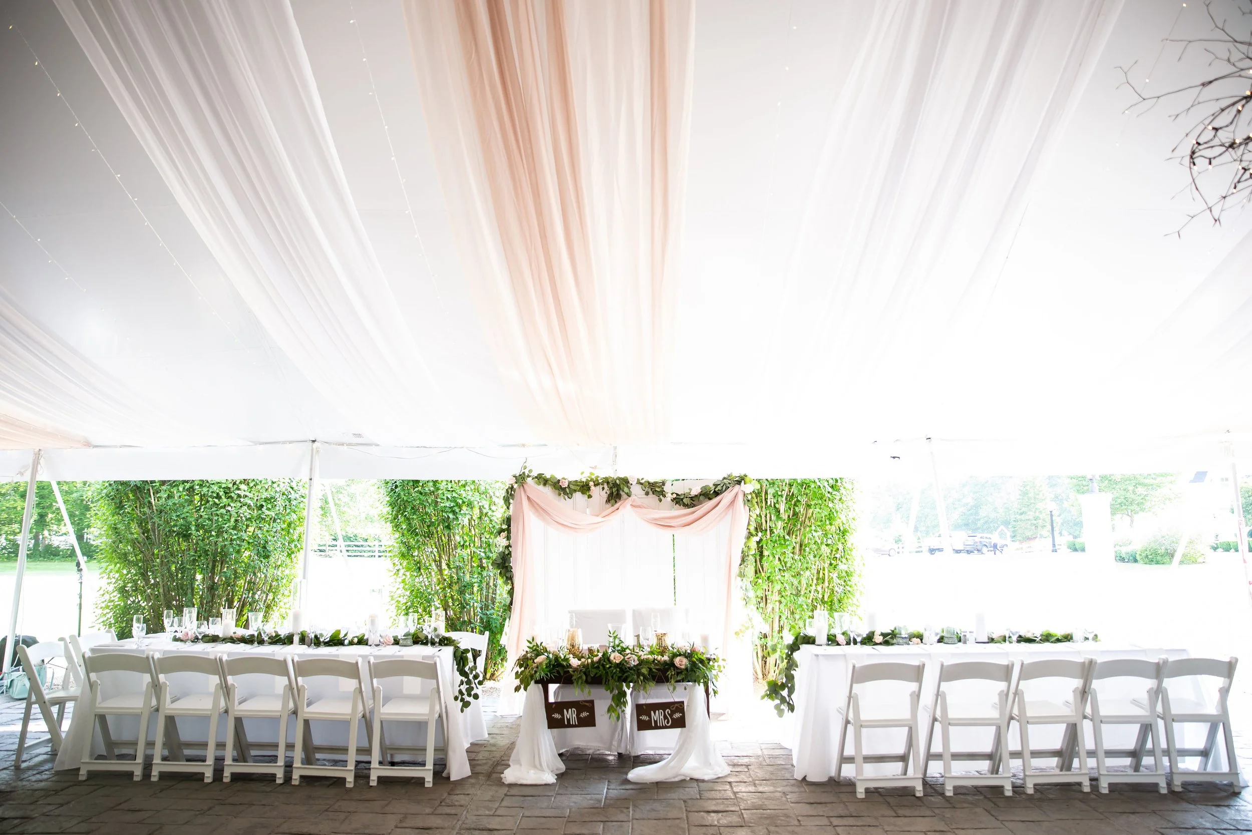 Wedding reception setup with a long table and chairs under a large white tent. The table is decorated with greenery and flowers, with a backdrop of greenery and pink drapery in the center.