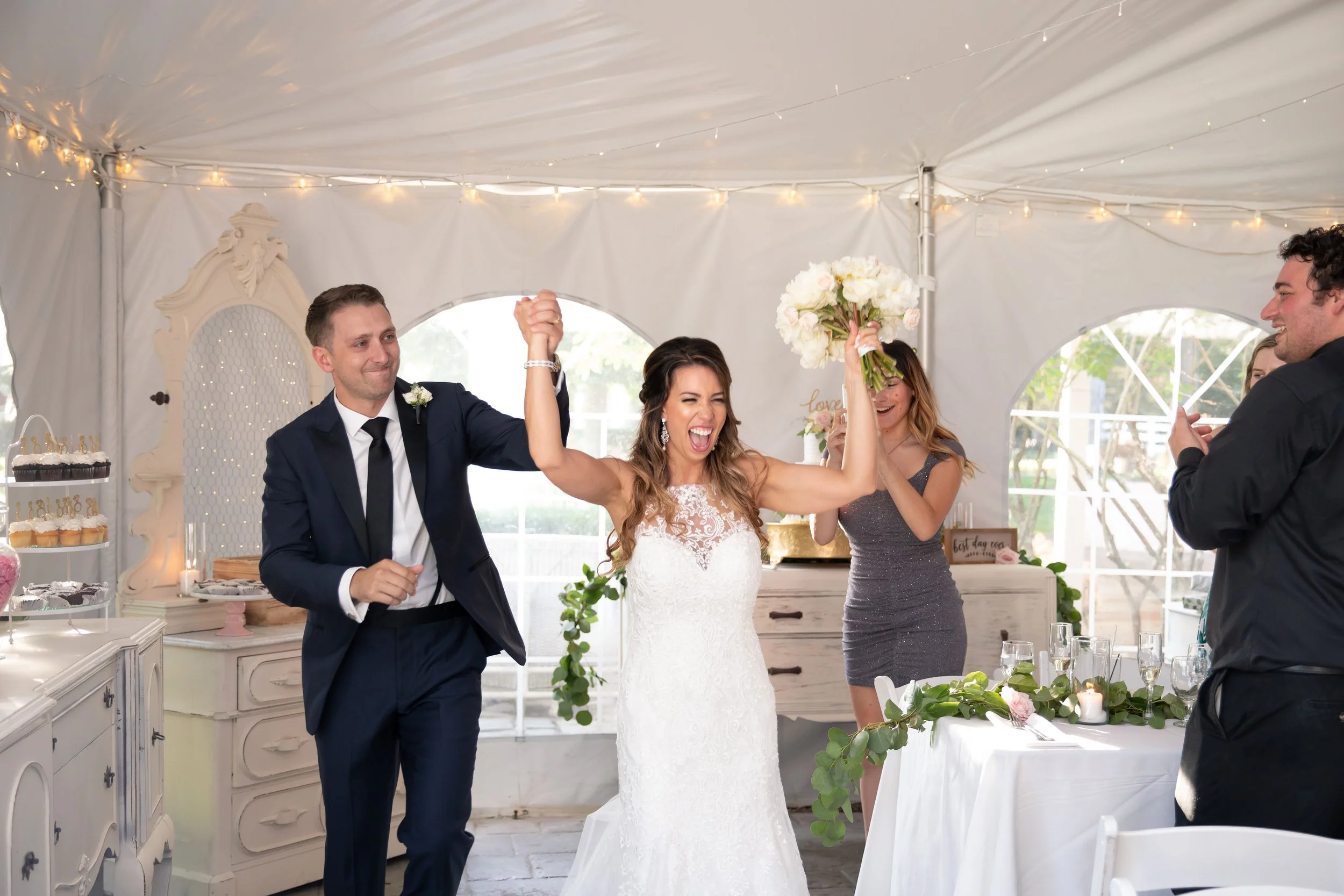 A bride in a white wedding dress celebrating with a man in a dark suit, holding hands and smiling at their wedding reception. Two women in the background are clapping and taking photos. The setting is decorated with flowers, candles, and fairy lights