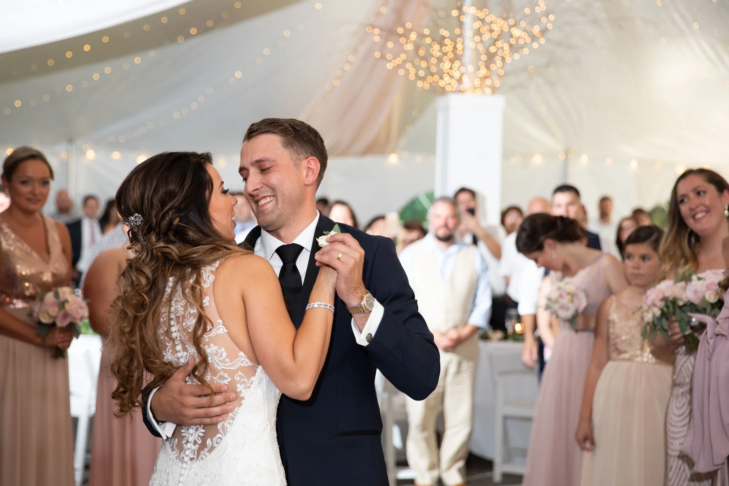 A bride and groom are dancing closely at their wedding reception under a decorated tent, surrounded by guests watching and smiling.