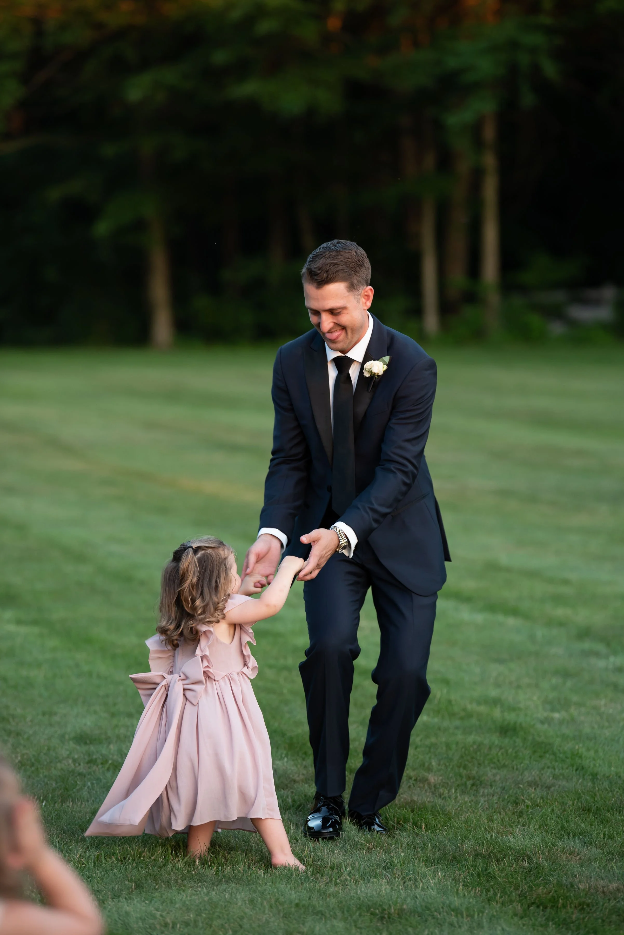 A groom in a black tuxedo dancing with a young girl in a pink dress at an outdoor wedding reception.