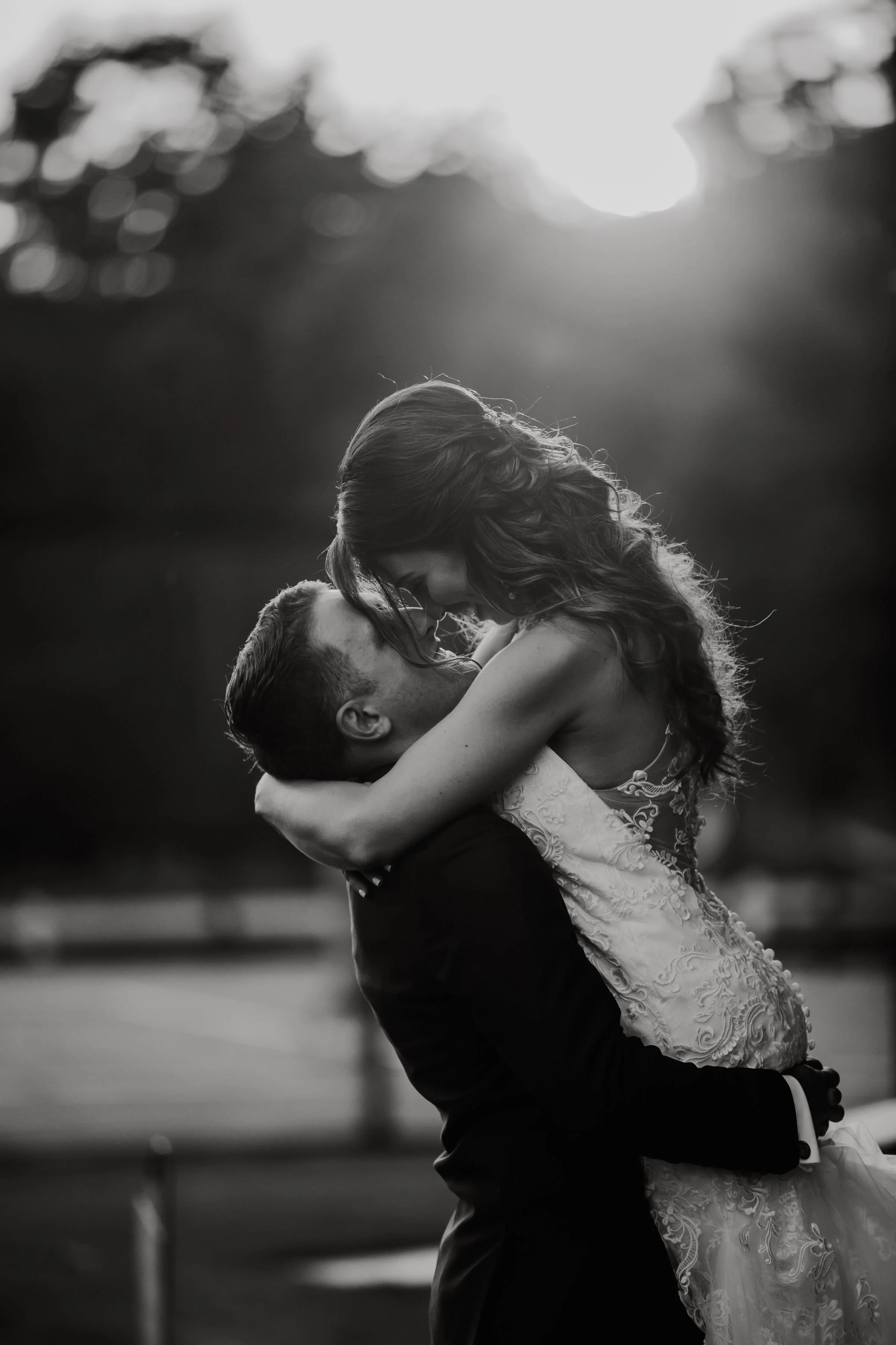 A black and white photo of a couple embracing, with the woman being lifted by the man during sunset or sunrise.