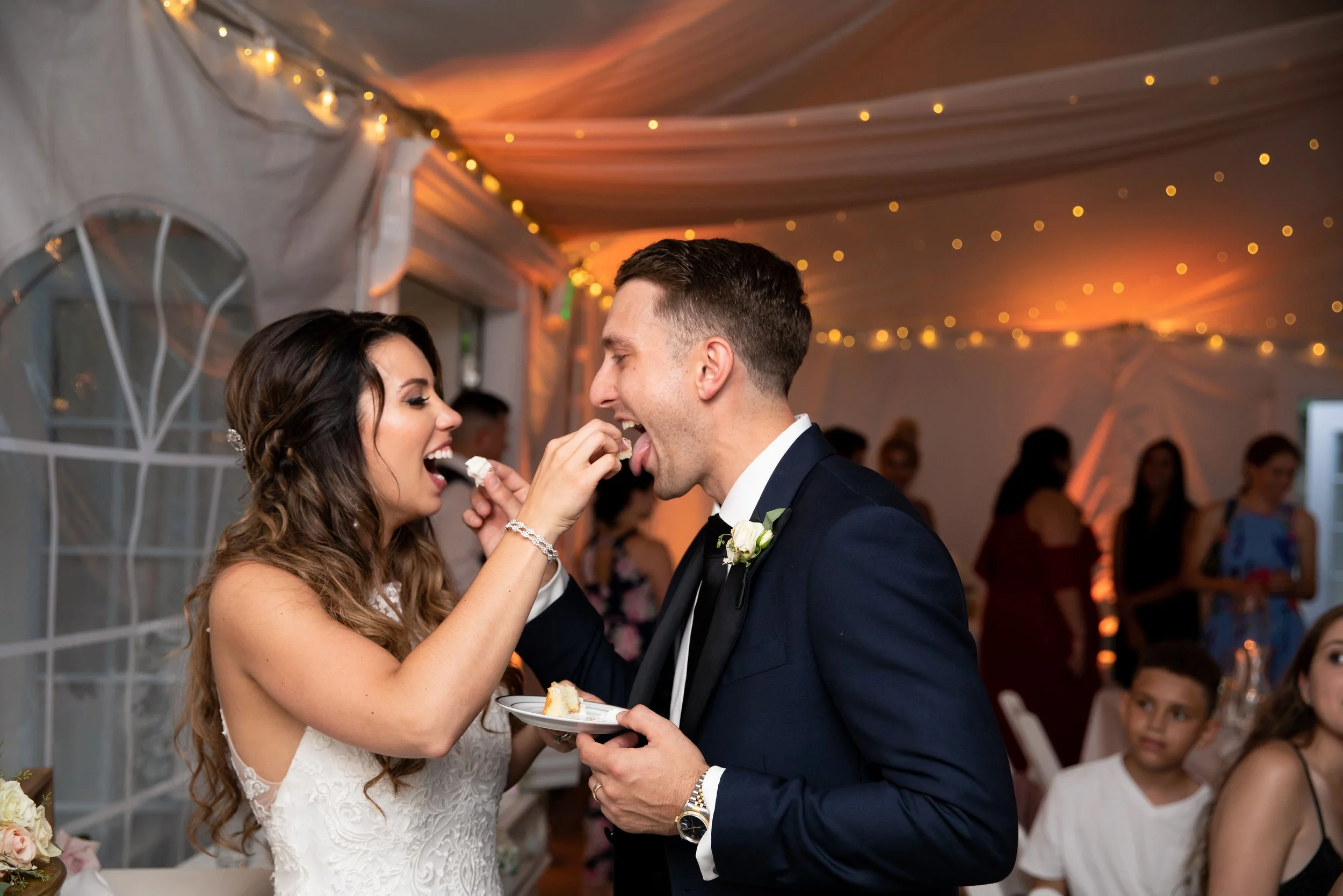 Bride and groom sharing cake at wedding reception in decorated tent with string lights and guests watching.