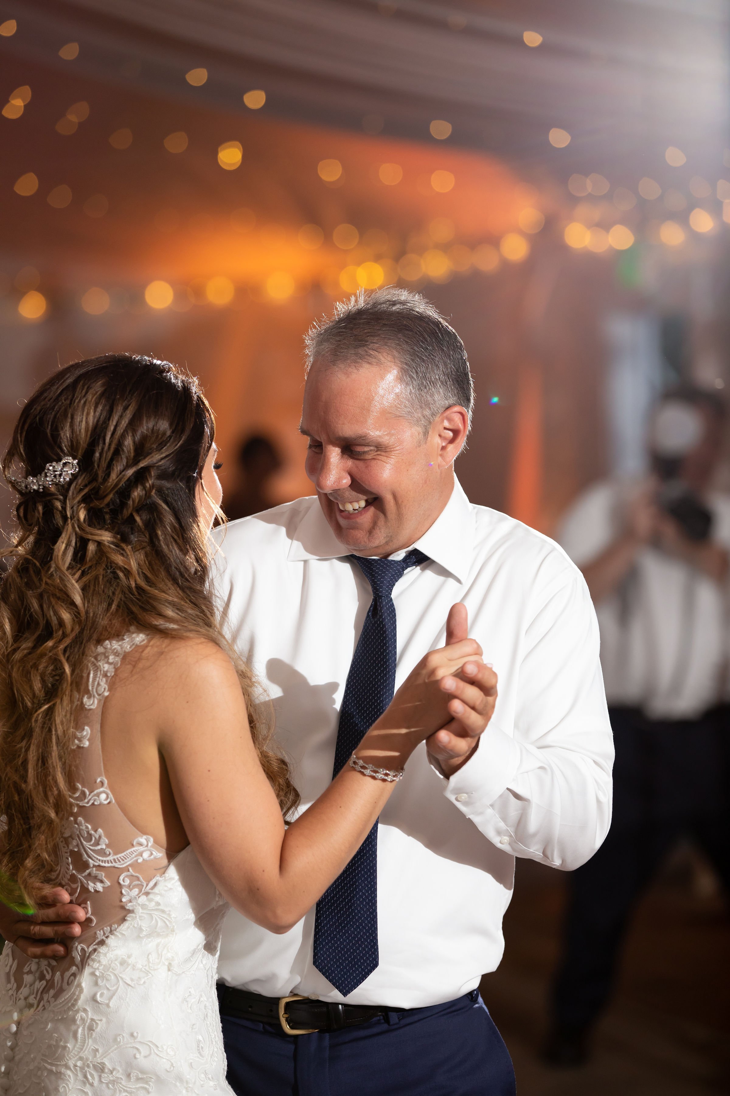A man and woman dancing at a wedding reception, with the man smiling and the woman wearing a lace wedding dress, in a warmly lit venue.