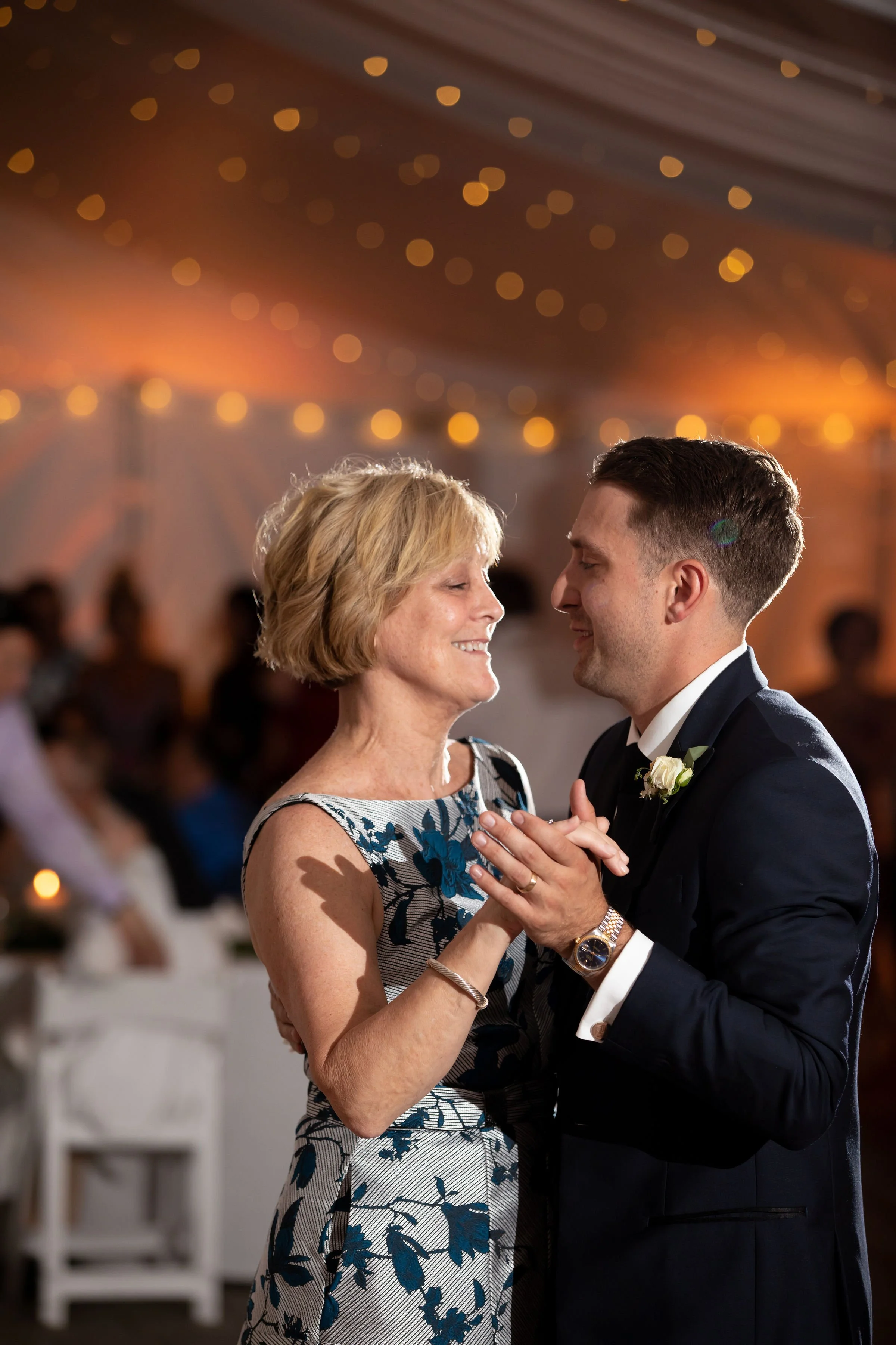 A woman and a man dancing closely at a wedding reception under warm, string lights. The woman is wearing a sleeveless floral dress, and the man is in a dark suit with a white shirt and boutonniere.