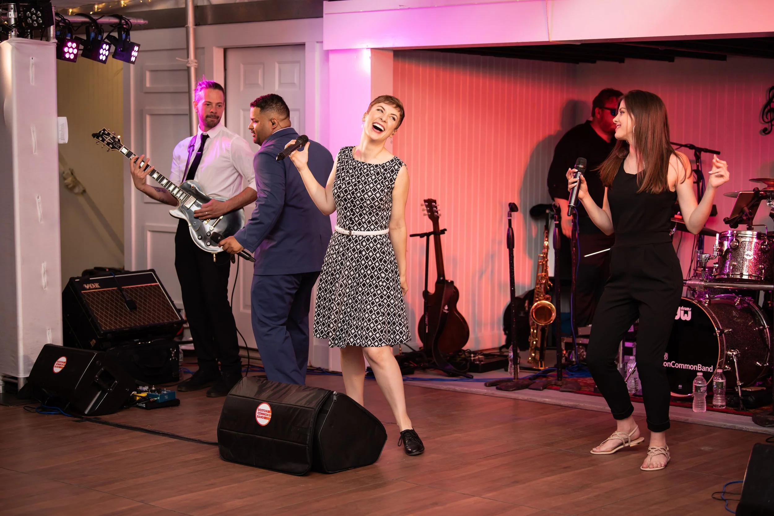 A band of five musicians performing on stage with two singers dancing and singing in front of the band. One singer is a woman in a black and white patterned dress, and the other is a woman in a black jumpsuit. The band includes a guitarist, bassist, 
