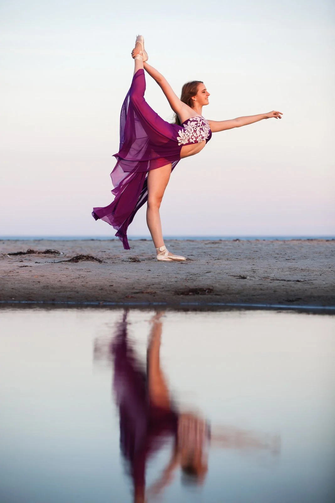 A woman in a purple dress practicing ballet on a sandy beach near water at sunset, with her reflection visible in the water.