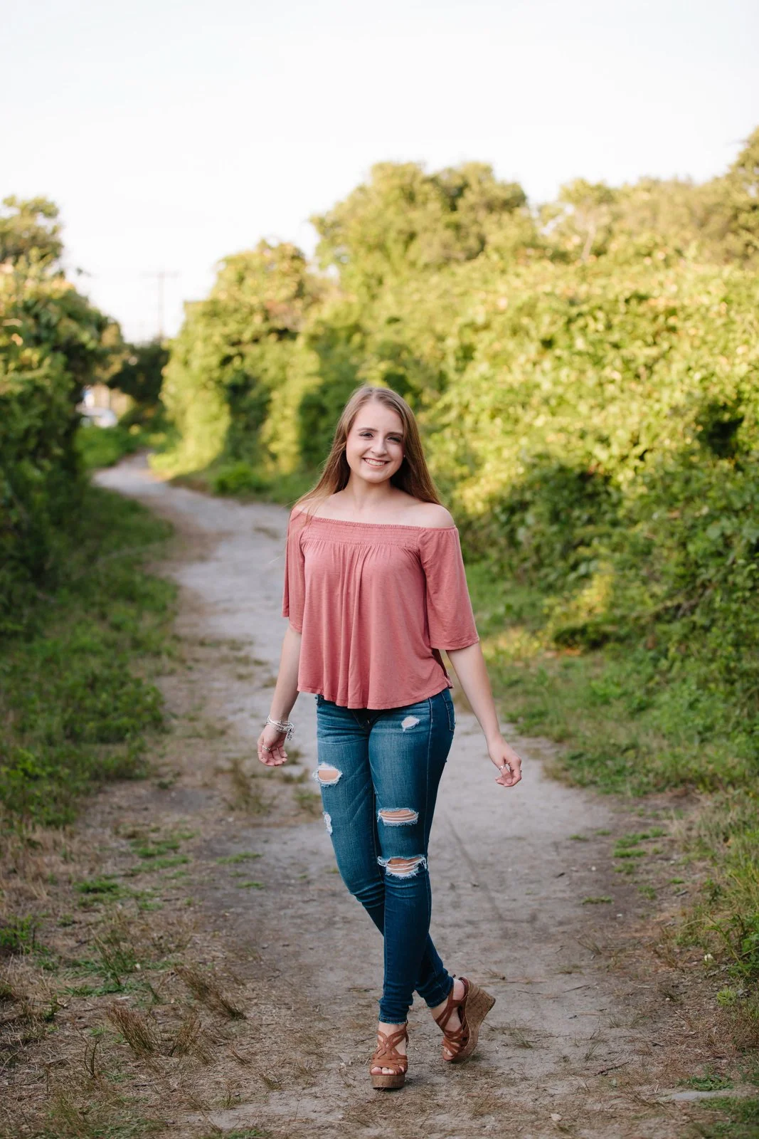 A young woman with long brown hair, wearing a pink off-the-shoulder top, ripped skinny jeans, and brown wedge sandals, walking on a dirt path surrounded by green bushes and trees, smiling at the camera.