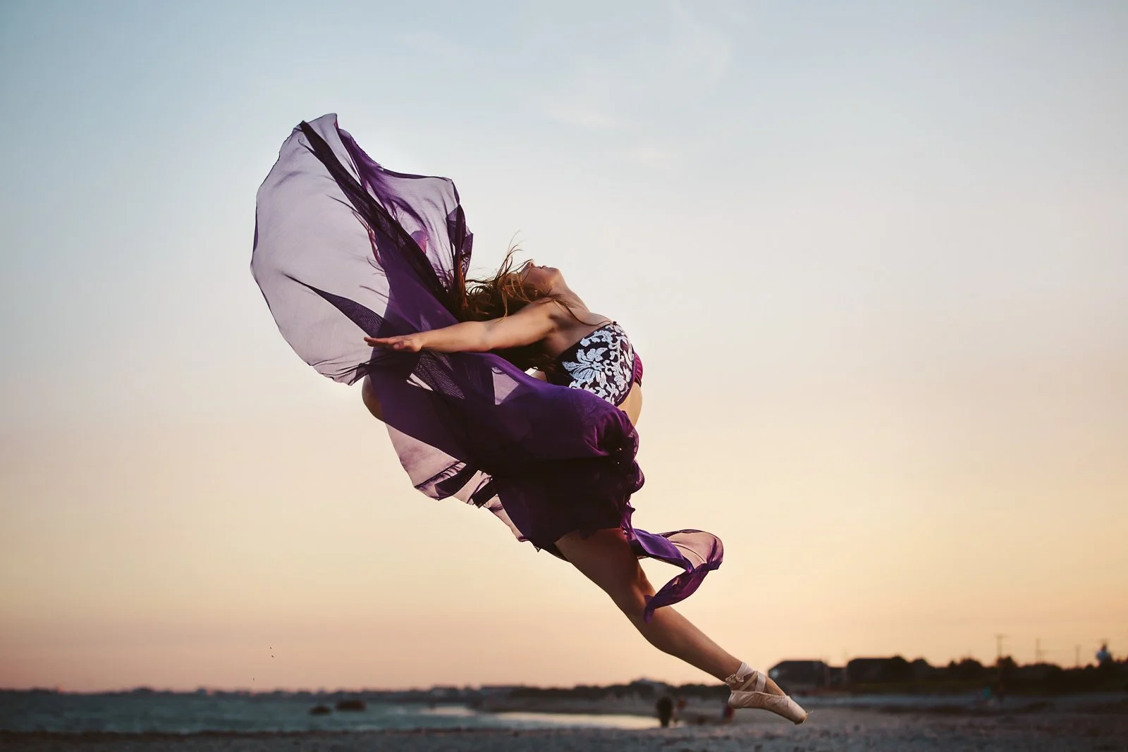 A dancer performing a leap on the beach during sunset, wearing a purple and black costume with flowing fabric.