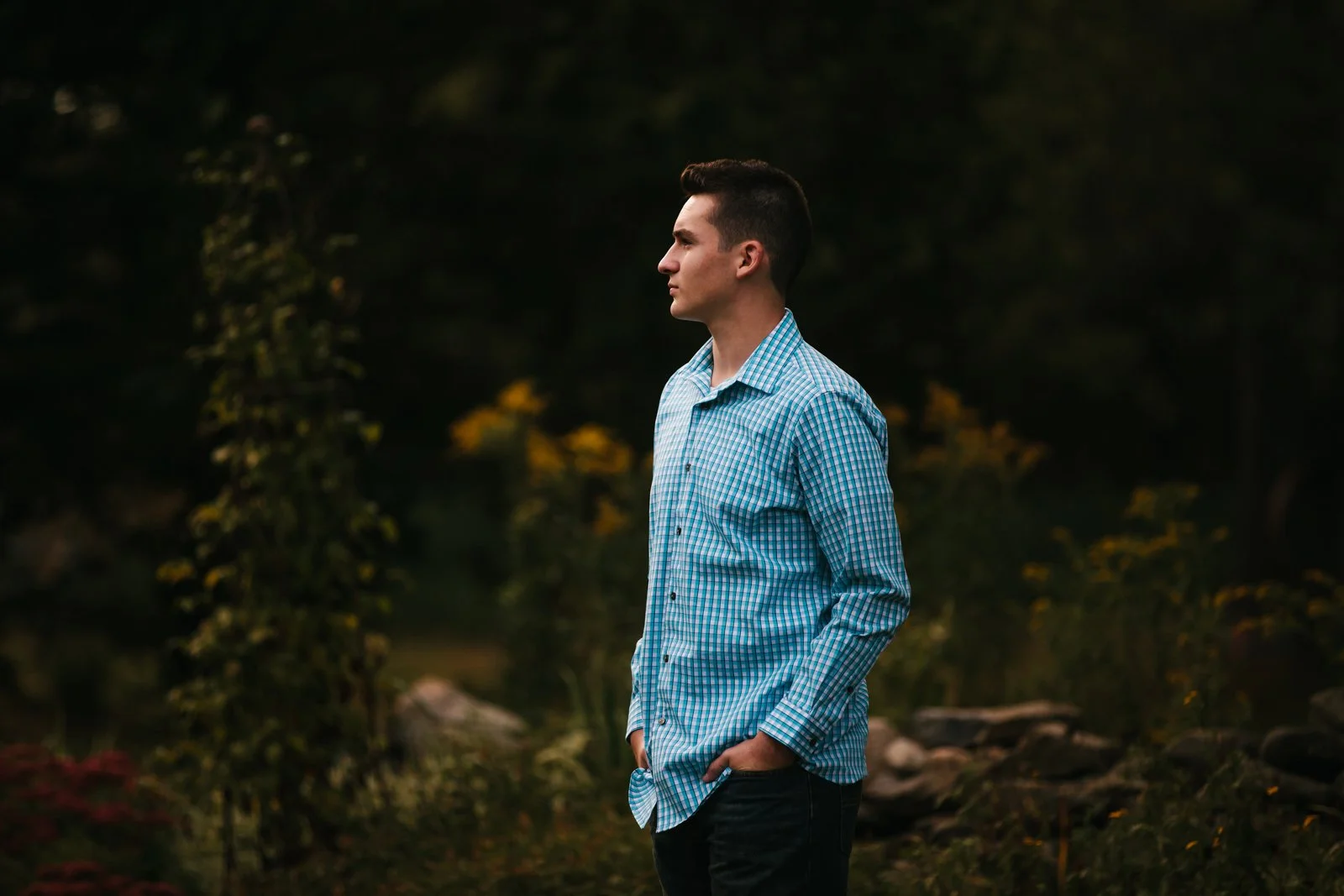 Young man standing outdoors in profile view, wearing a blue checkered shirt with hands in pockets, against a blurred natural background.