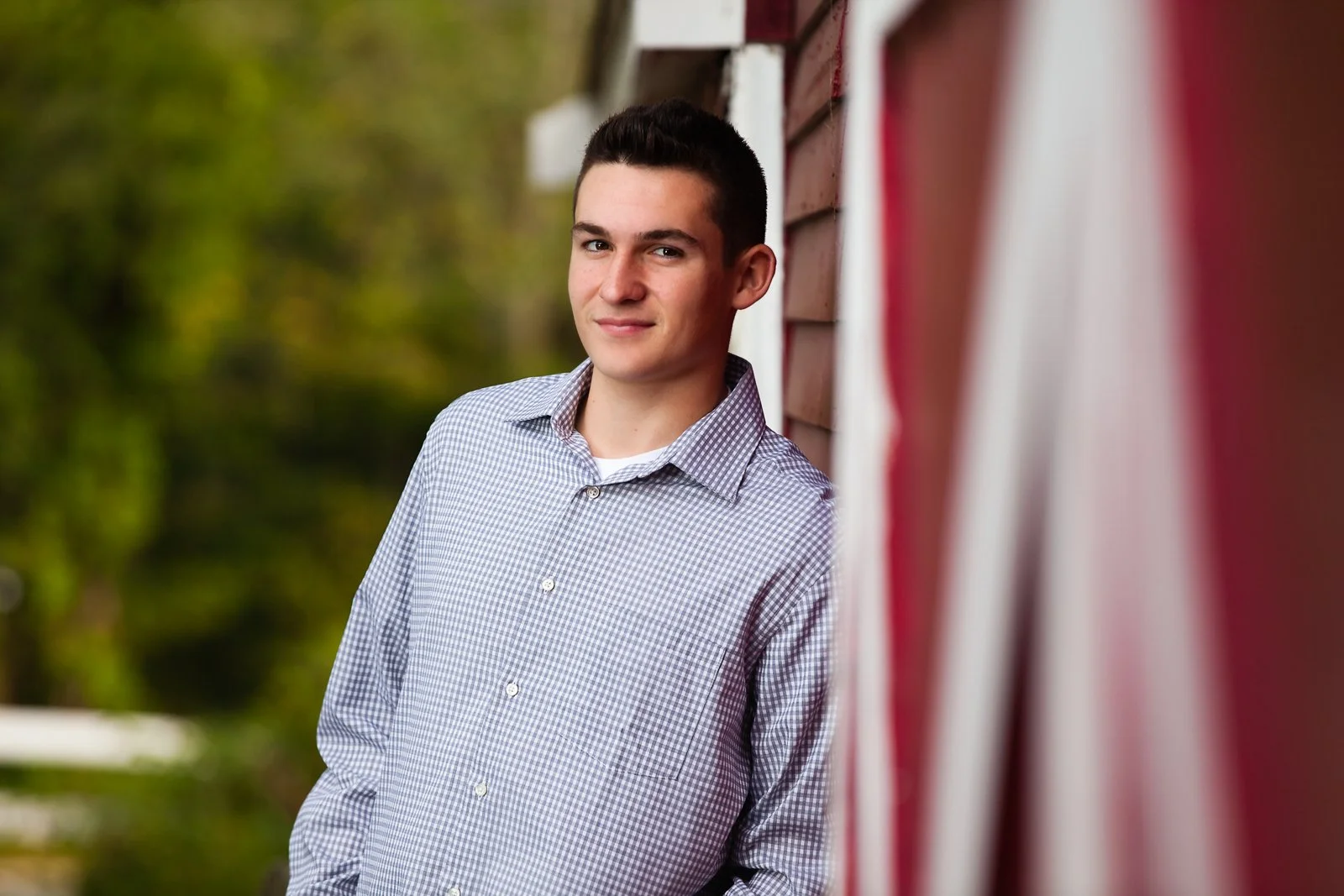 A young man with dark hair wearing a checked button-up shirt, standing outdoors next to a red building, smiling slightly, with greenery in the background.