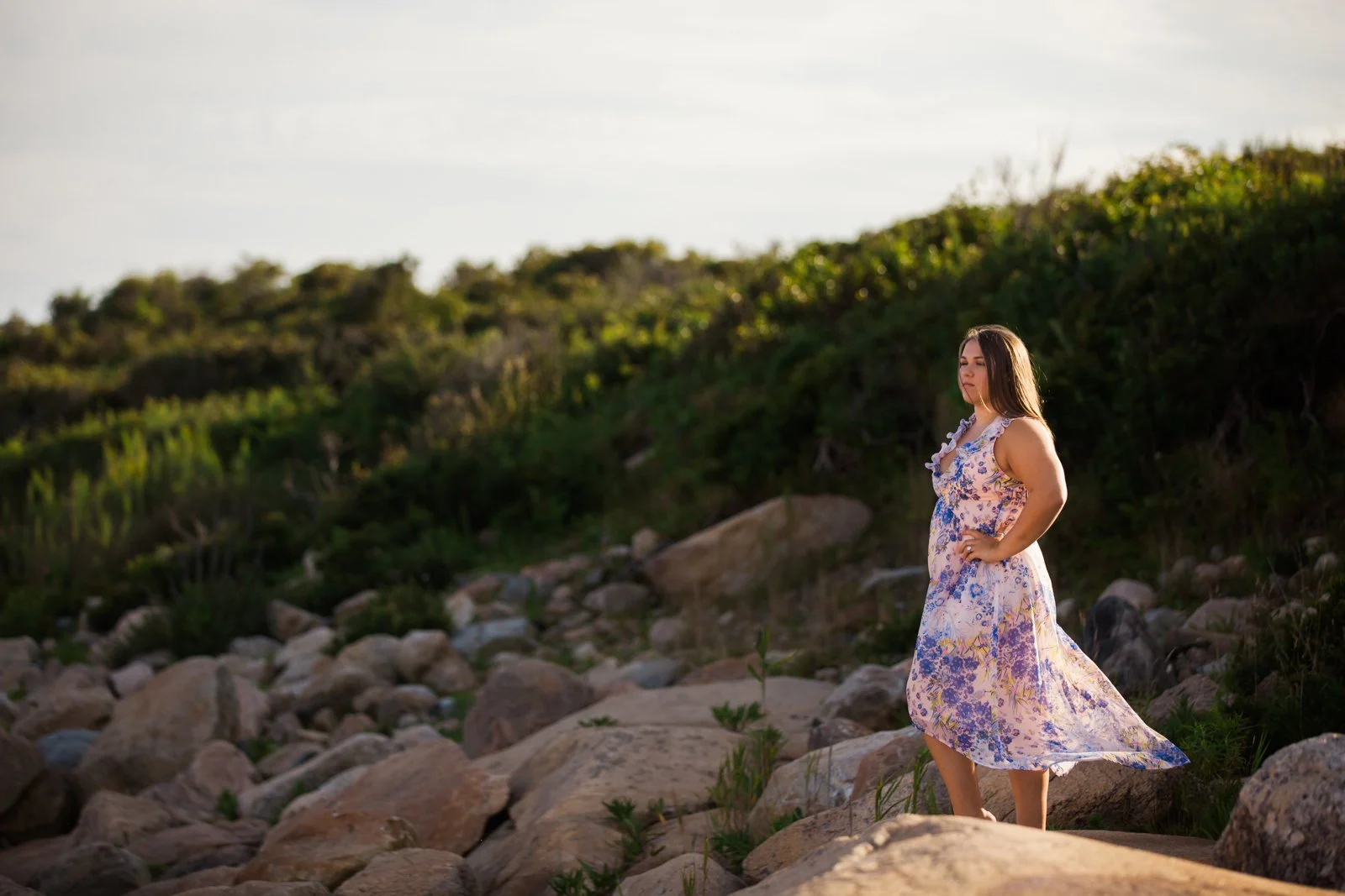 Woman in a floral dress standing on rocks in a natural outdoor setting with a green hillside in the background.