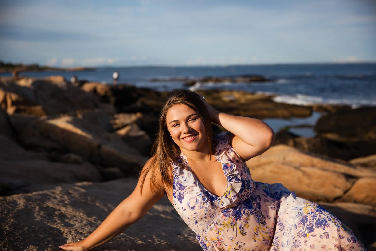A young woman smiling and sitting on rocks at the beach during sunset.