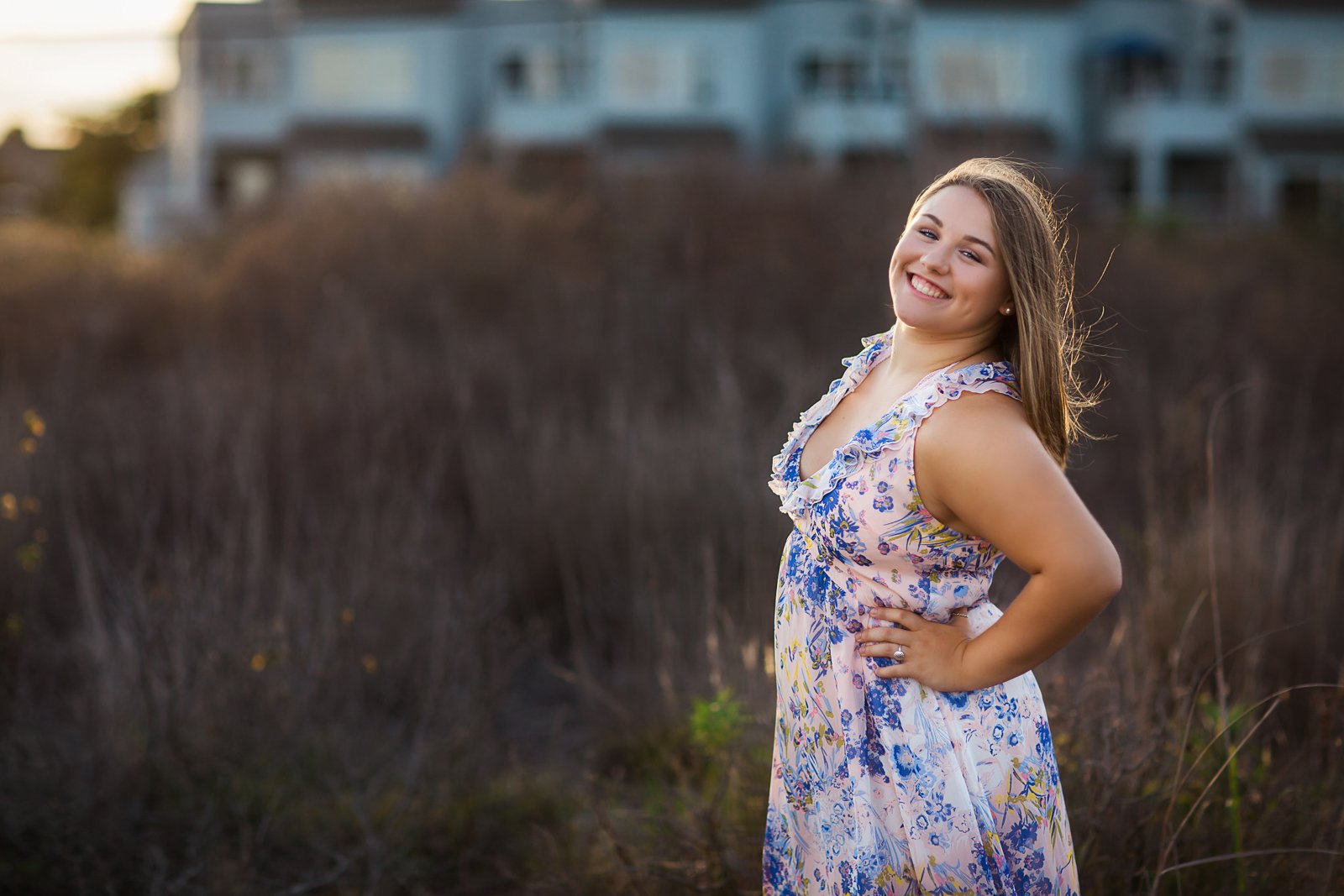 A young woman with long brown hair, wearing a sleeveless floral dress, smiling and posing outdoors in front of dry bushes and a building in the background.