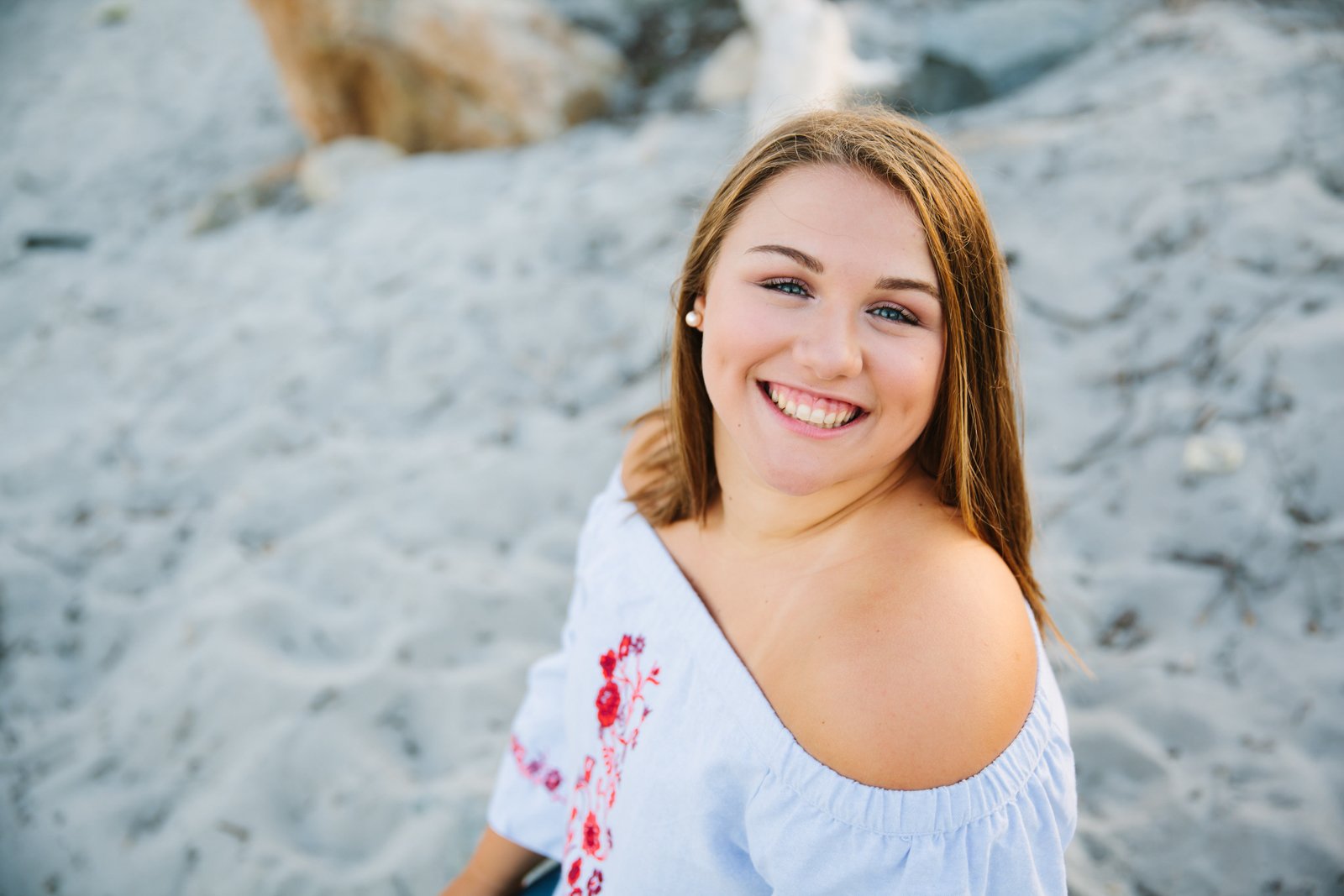 A young woman with long, light brown hair smiling at the camera, sitting on a sandy beach with rocks in the background, wearing a white off-shoulder top with red floral embroidery.