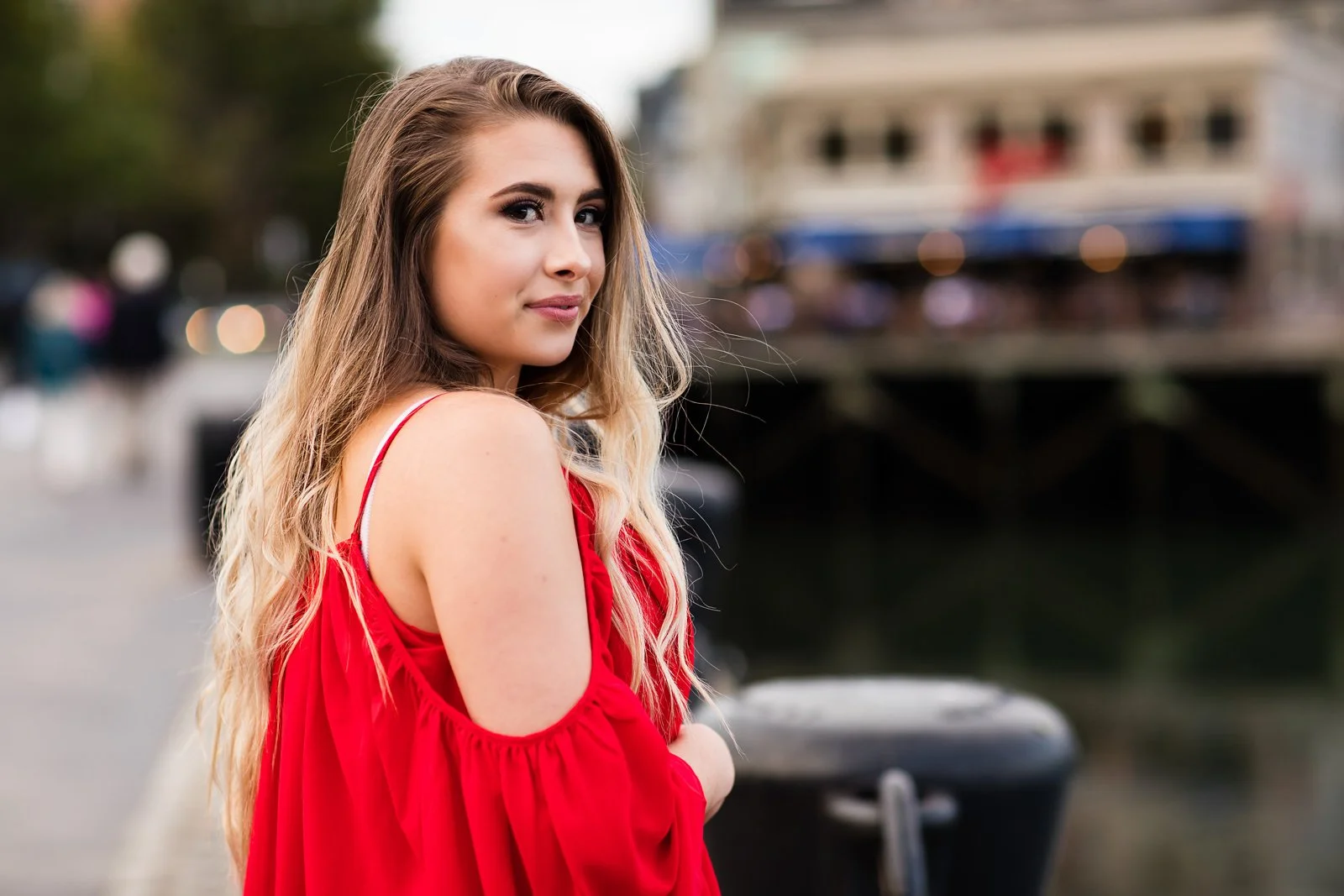 A young woman with long blond hair in a red cold shoulder dress standing outdoors near a body of water with blurred people and buildings in the background.