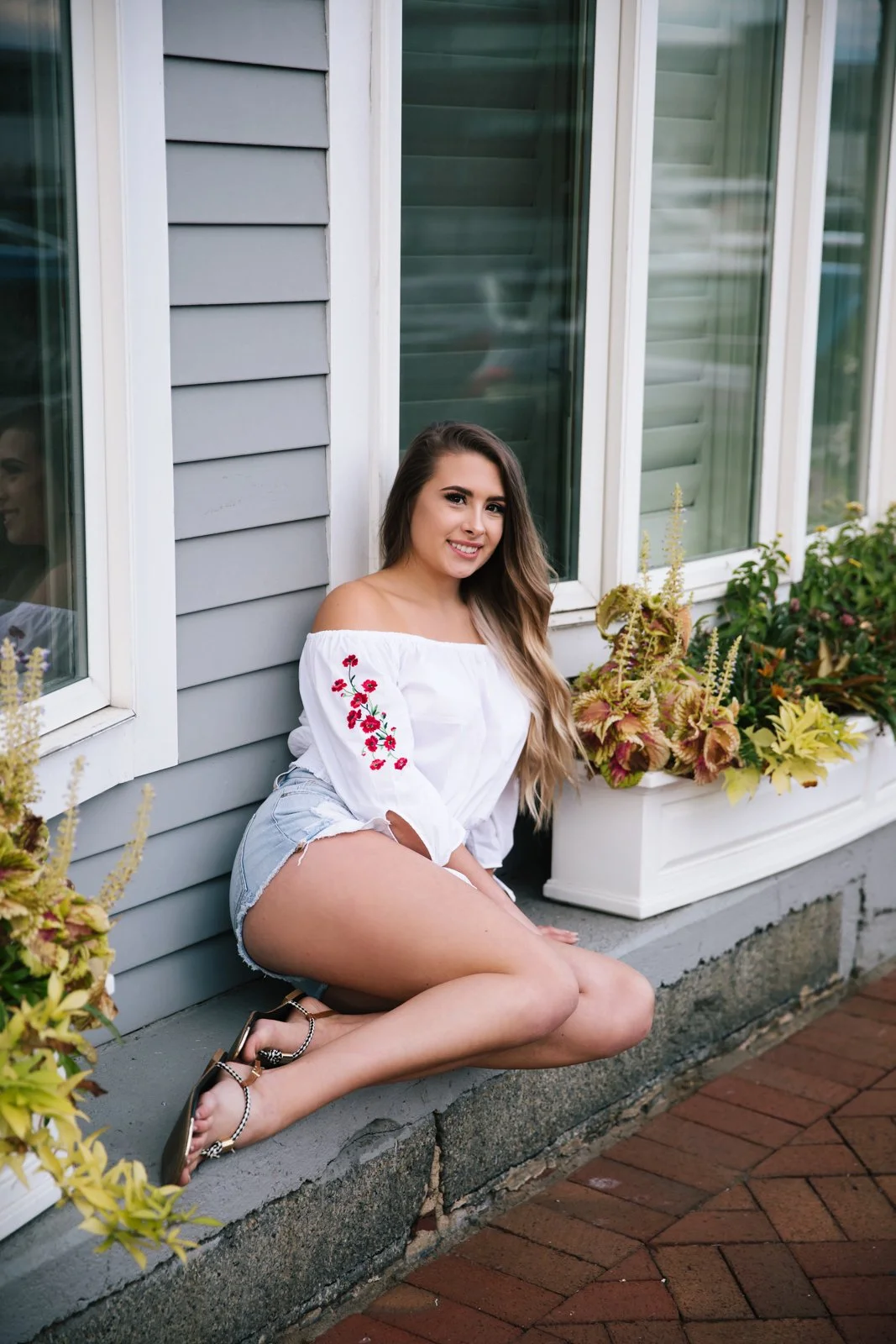 A young woman with long, wavy hair, wearing a white off-shoulder blouse with red floral embroidery, denim shorts, and sandals, sitting on a concrete ledge beside a house with gray siding and large windows, next to potted plants with colorful foliage.