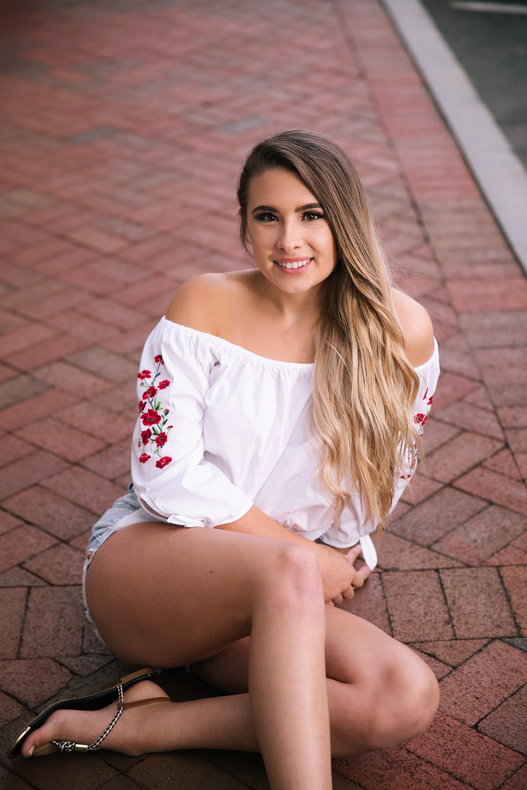 A young woman with long wavy hair sitting on a brick sidewalk, smiling at the camera, wearing an off-shoulder white top with red embroidery, denim shorts, and black sandals.